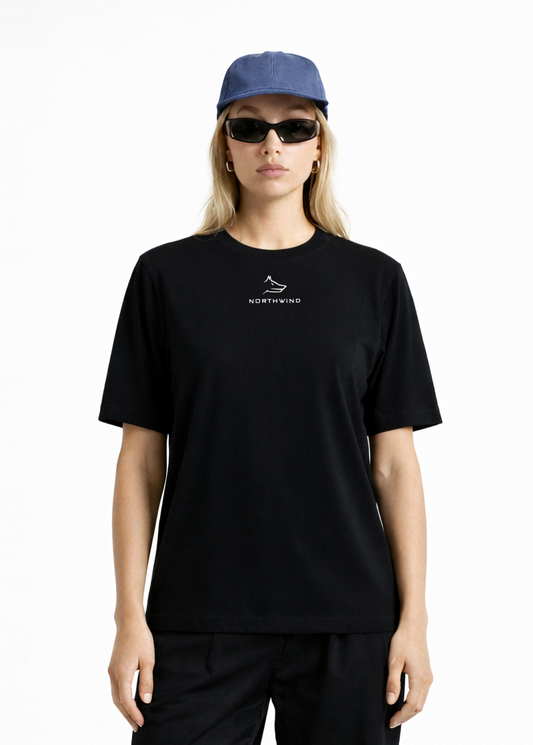Woman wearing a black Northwind casual t-shirt with minimalist Northwind embroidered wolf logo on the chest, blue hat, black sunglasses and black pants on a white background