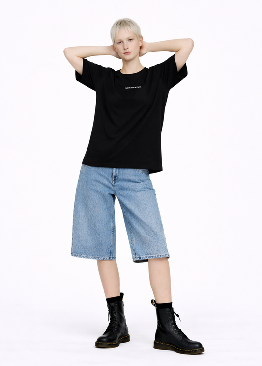 Blond, short hair girl wearing a black Northwind casual storm of wolves t-shirt with Northwind logo embroidered on the chest, blue denim Carhartt oversized shorts, black belt, and black Dr. Martens boots on a white background