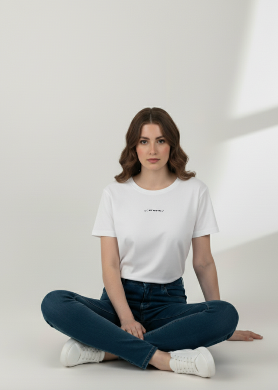 Woman sitting on the floor wearing Essential White T-shirt with Northwind Logo Embroidery and dark jeans in a minimalistic setting