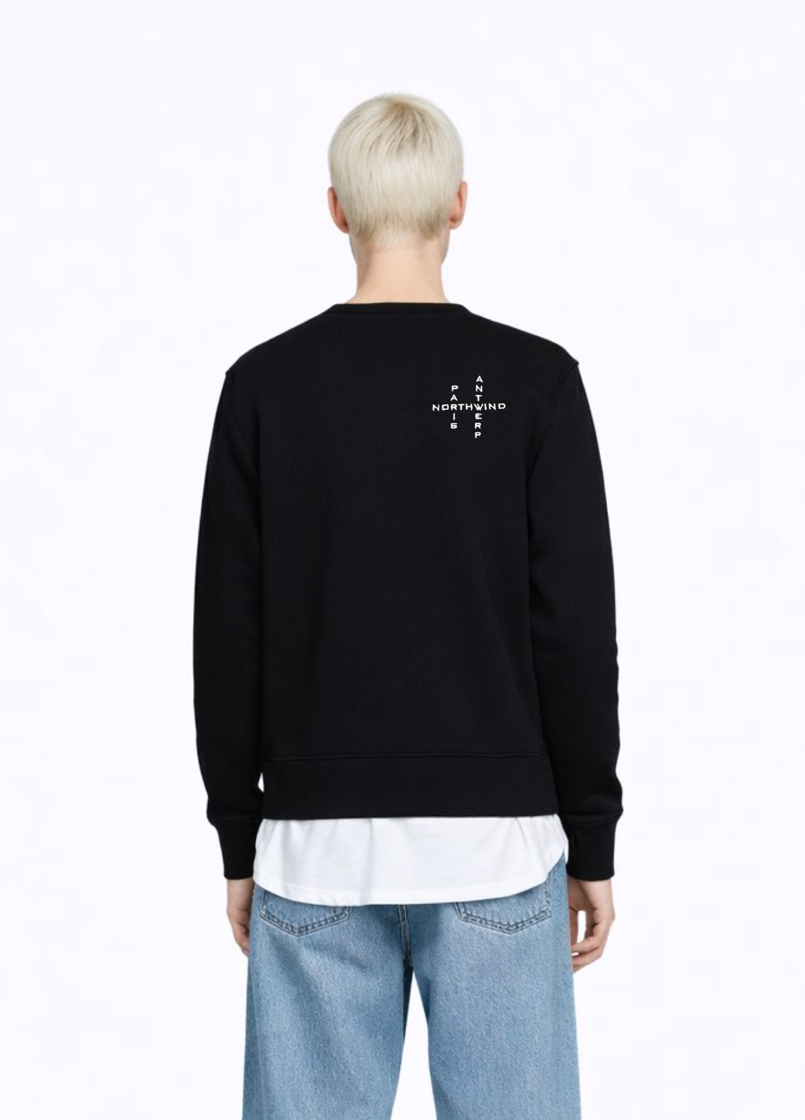 A blond, short hair woman wearing a black Northwind Crossword sweatshirt with Northwind crossword Antwerp Paris embroidered logo on the back, white Northwind t-shirt under the sweatshirt, blue jeans