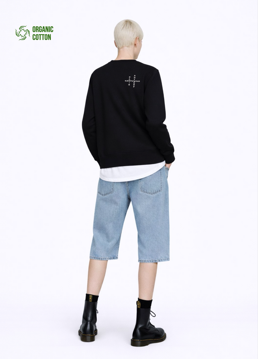 A blond, short hair woman wearing a black organic cotton Northwind Crossword sweatshirt with Northwind crossword Antwerp Paris embroidered logo on the back, white Northwind t-shirt under the sweatshirt, blue jeans, and black Dr Martens boots