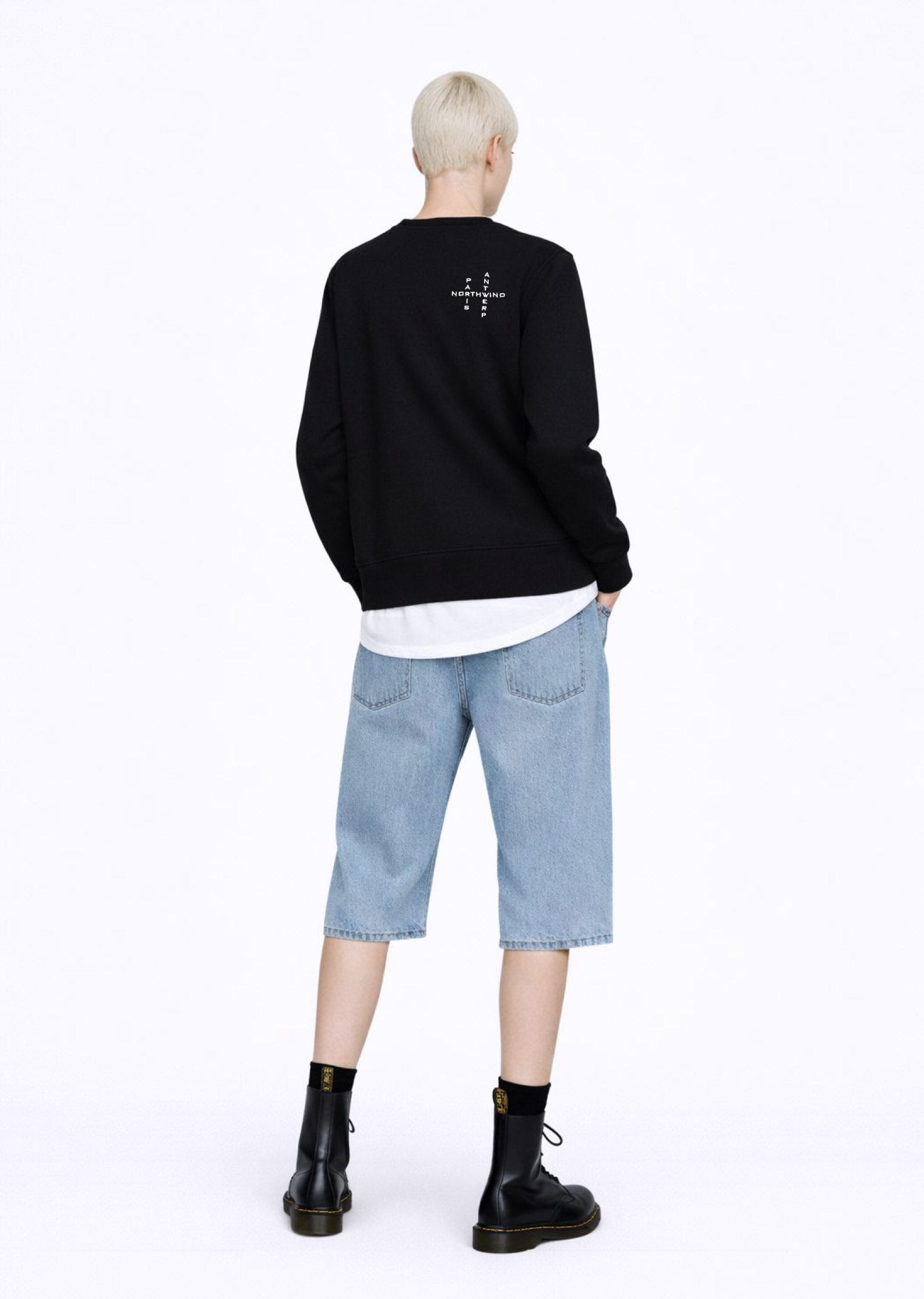 A blond, short hair woman wearing a black Northwind Crossword sweatshirt with Northwind crossword Antwerp Paris embroidered logo on the back, white Northwind t-shirt under the sweatshirt, blue jeans, and black Dr Martens boots