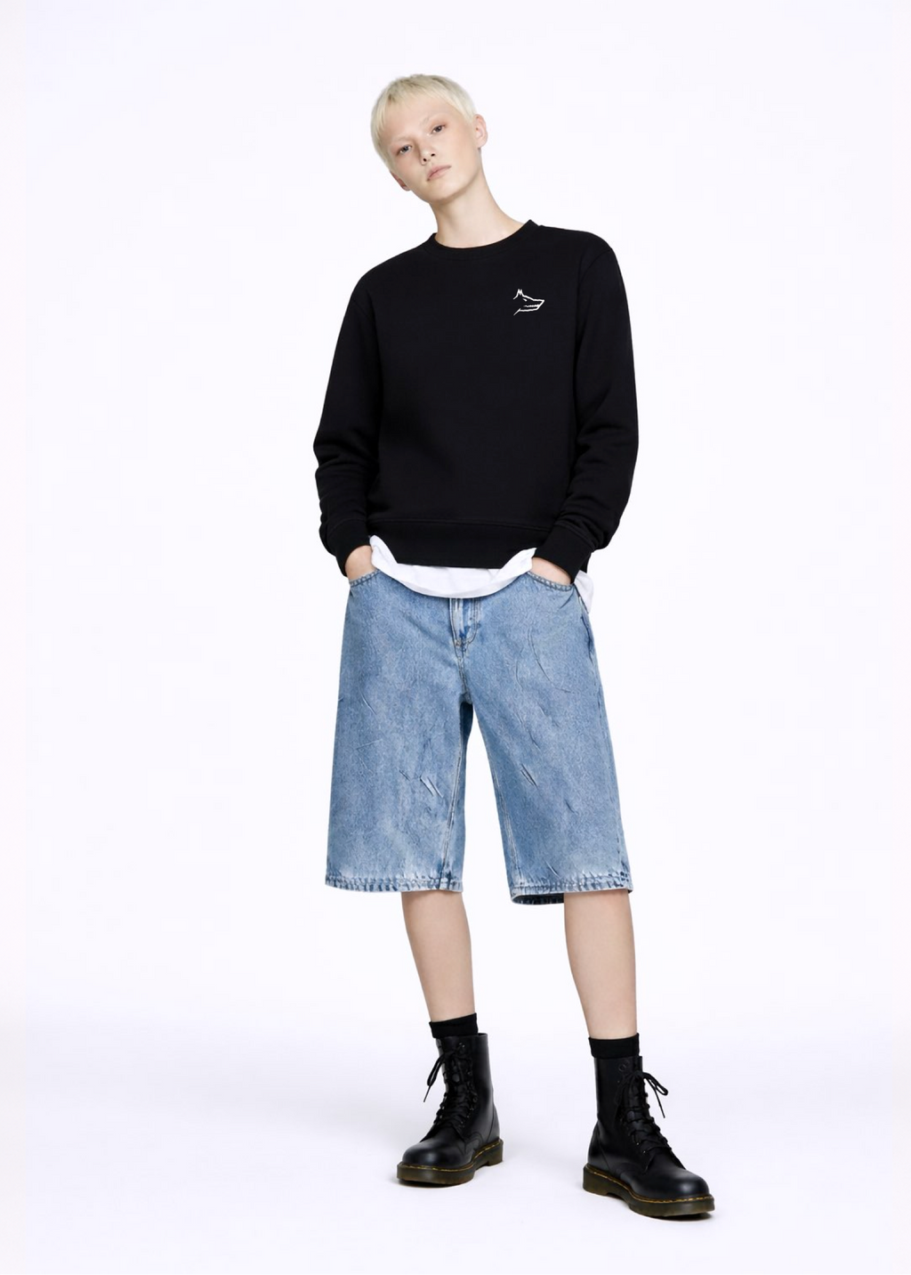 A blond, short hair woman wearing a black Northwind Crossword sweatshirt with Northwind wolf embroidered logo on the chest, white Northwind t-shirt under the sweatshirt, blue jeans, and black Dr Martens boots