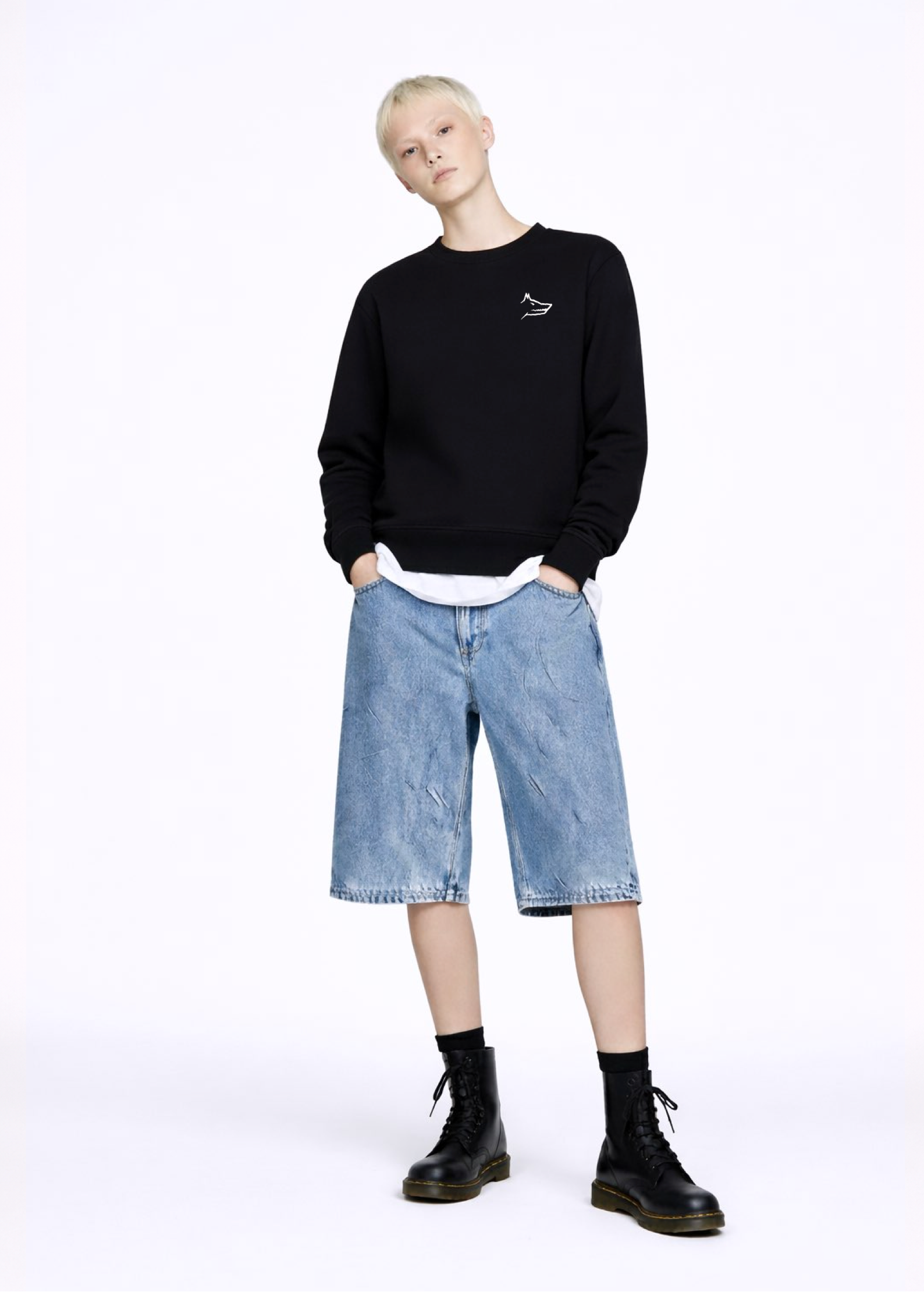 A blond, short hair woman wearing a black Northwind Crossword sweatshirt with Northwind wolf embroidered logo on the chest, white Northwind t-shirt under the sweatshirt, blue jeans, and black Dr Martens boots