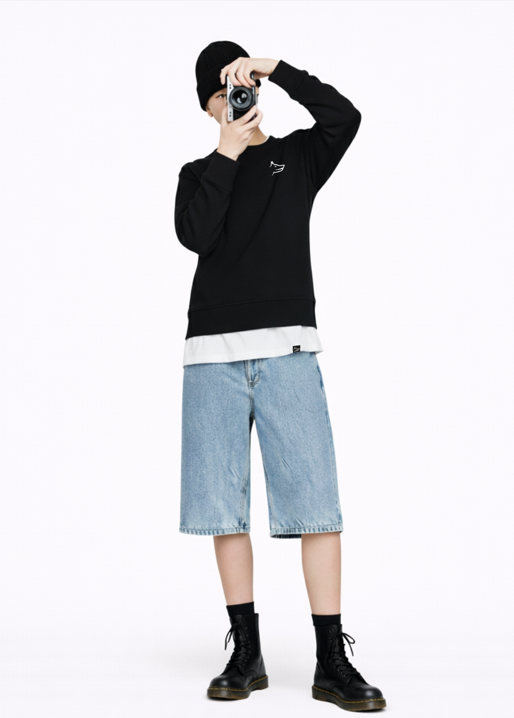 Woman wearing a black Northwind Crossword sweatshirt with Northwind wolf embroidered logo on the chest, Northwind black organic beanie, white shirt, blue jeans, and black Dr Martens boots and holding a camera on her hand