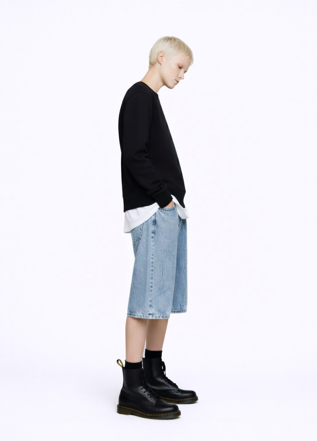 A blond, short hair woman wearing a black Northwind Crossword sweatshirt with Northwind crossword Antwerp Paris embroidered logo on the back, white Northwind t-shirt under the sweatshirt, blue jeans, and black Dr Martens boots