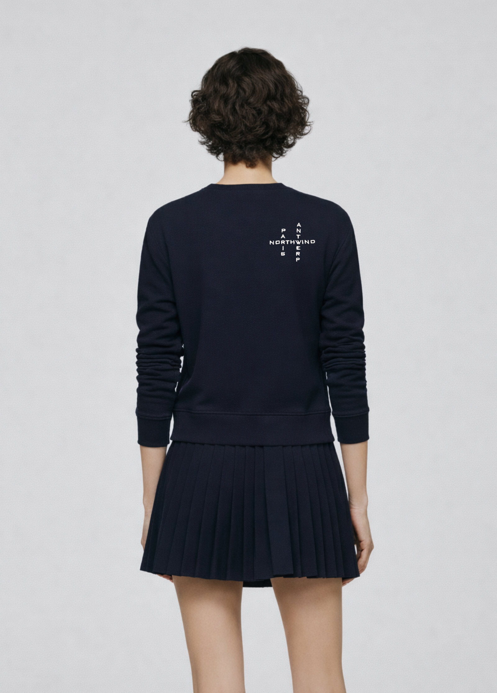 Woman wearing a french navy blue Northwind sweatshirt with Northwind crossword Antwerp Paris embroidered logo on the back, navy skirt