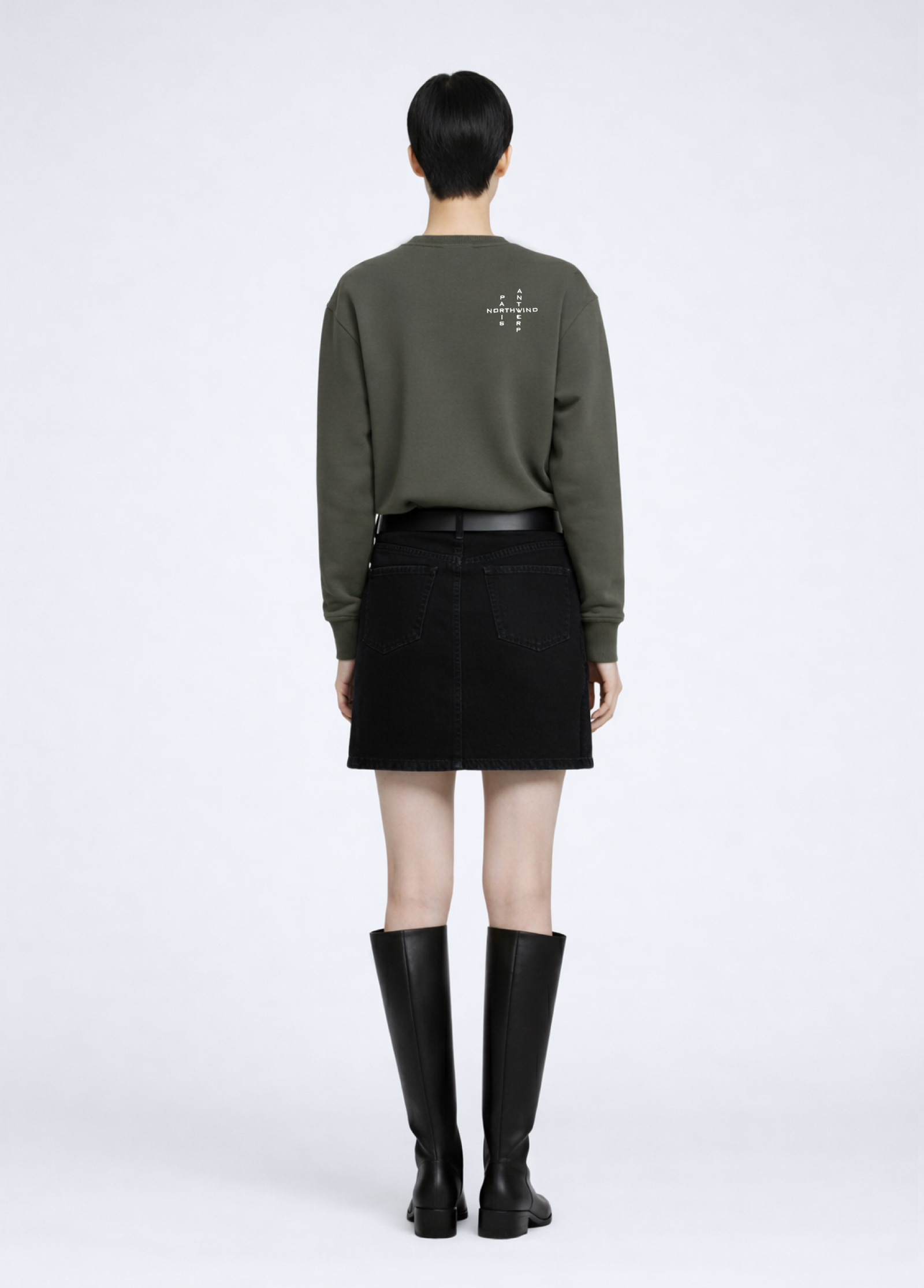 woman wearing a khaki green Northwind Crossword sweatshirt with Crossword Antwerp Paris embroidered logo on the back, black skirt, and black knee-high boots on a white background