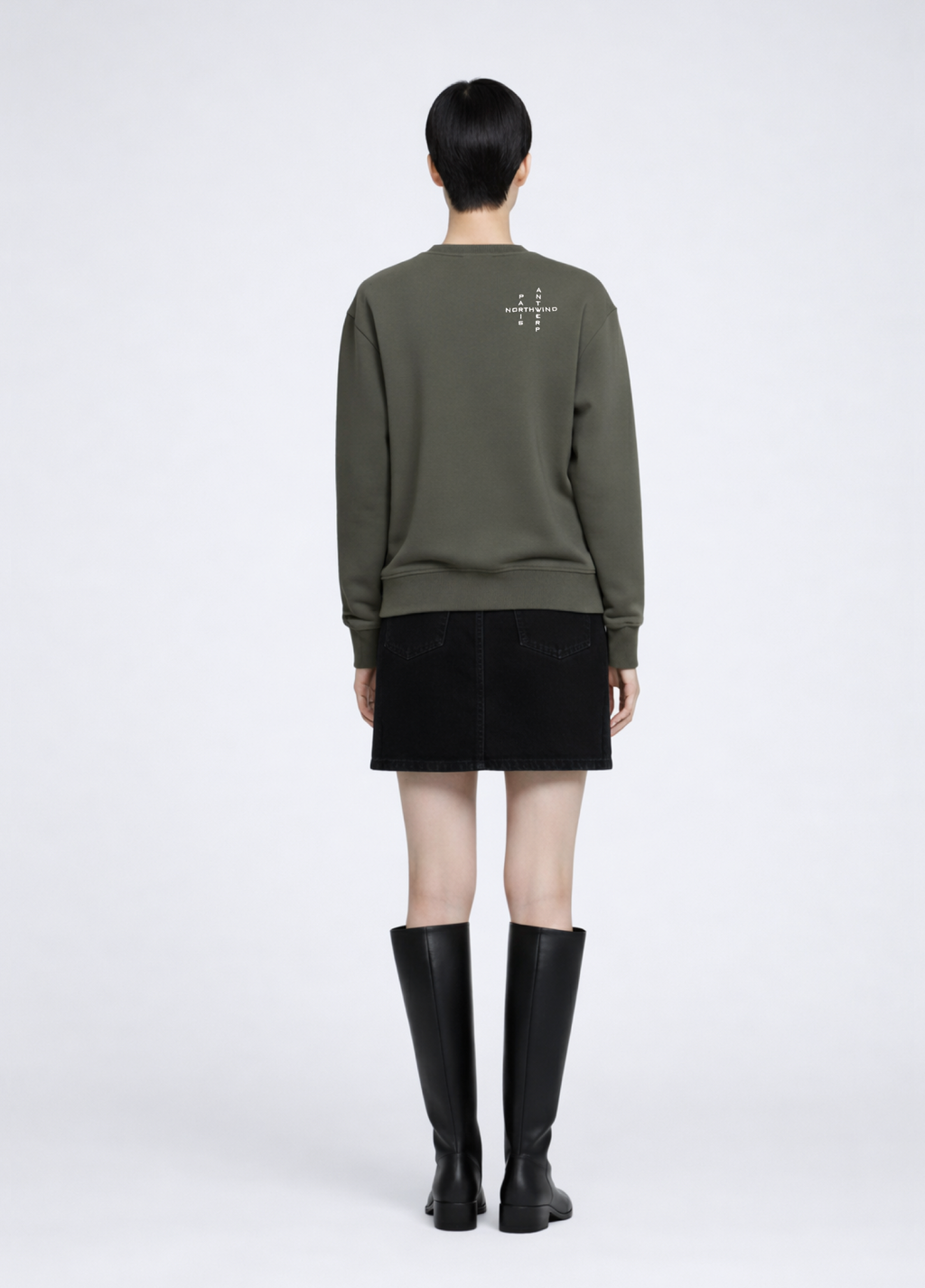 woman wearing a khaki green Northwind Crossword sweatshirt with Crossword Antwerp Paris embroidered logo on the back, black skirt, and black knee-high boots on a white background