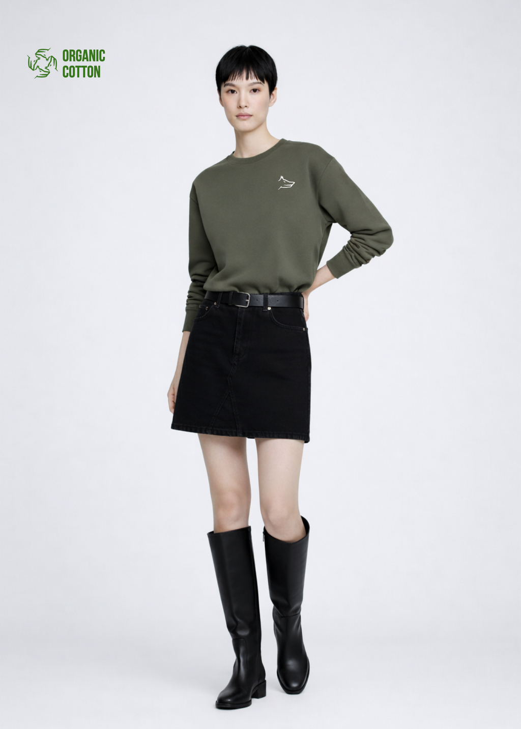 Asian woman wearing a khaki green organic cotton Northwind Crossword sweatshirt with Northwind wolf embroidered logo on the chest, black skirt, and black knee-high boots on a white background