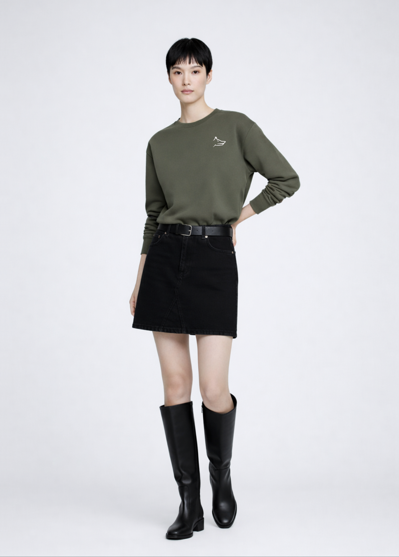 Asian woman wearing a khaki green Northwind Crossword sweatshirt with Northwind wolf embroidered logo on the chest, black skirt, and black knee-high boots on a white background