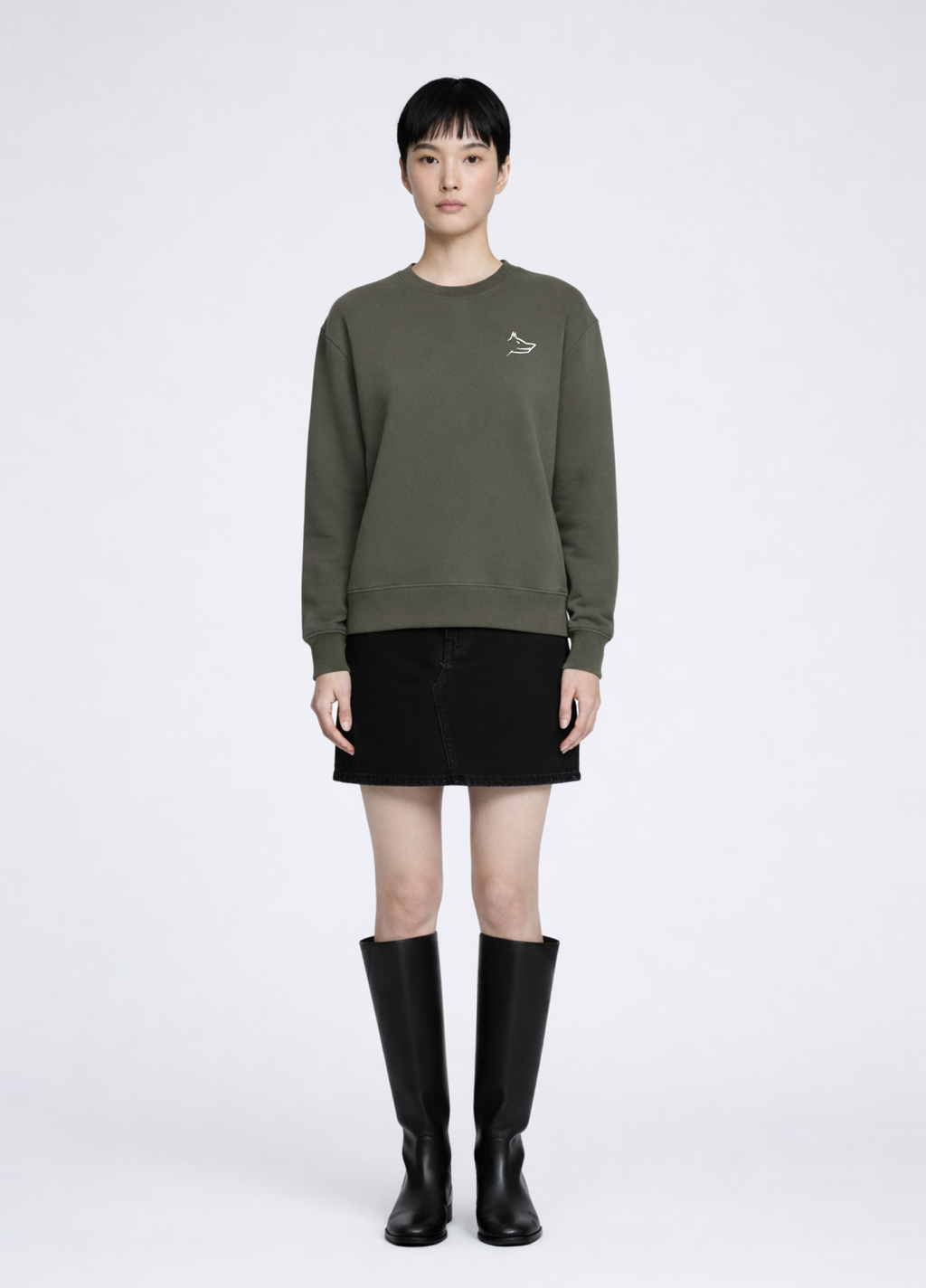 Asian woman wearing a khaki green Northwind Crossword sweatshirt with Northwind wolf embroidered logo on the chest, black skirt, and black knee-high boots on a white background