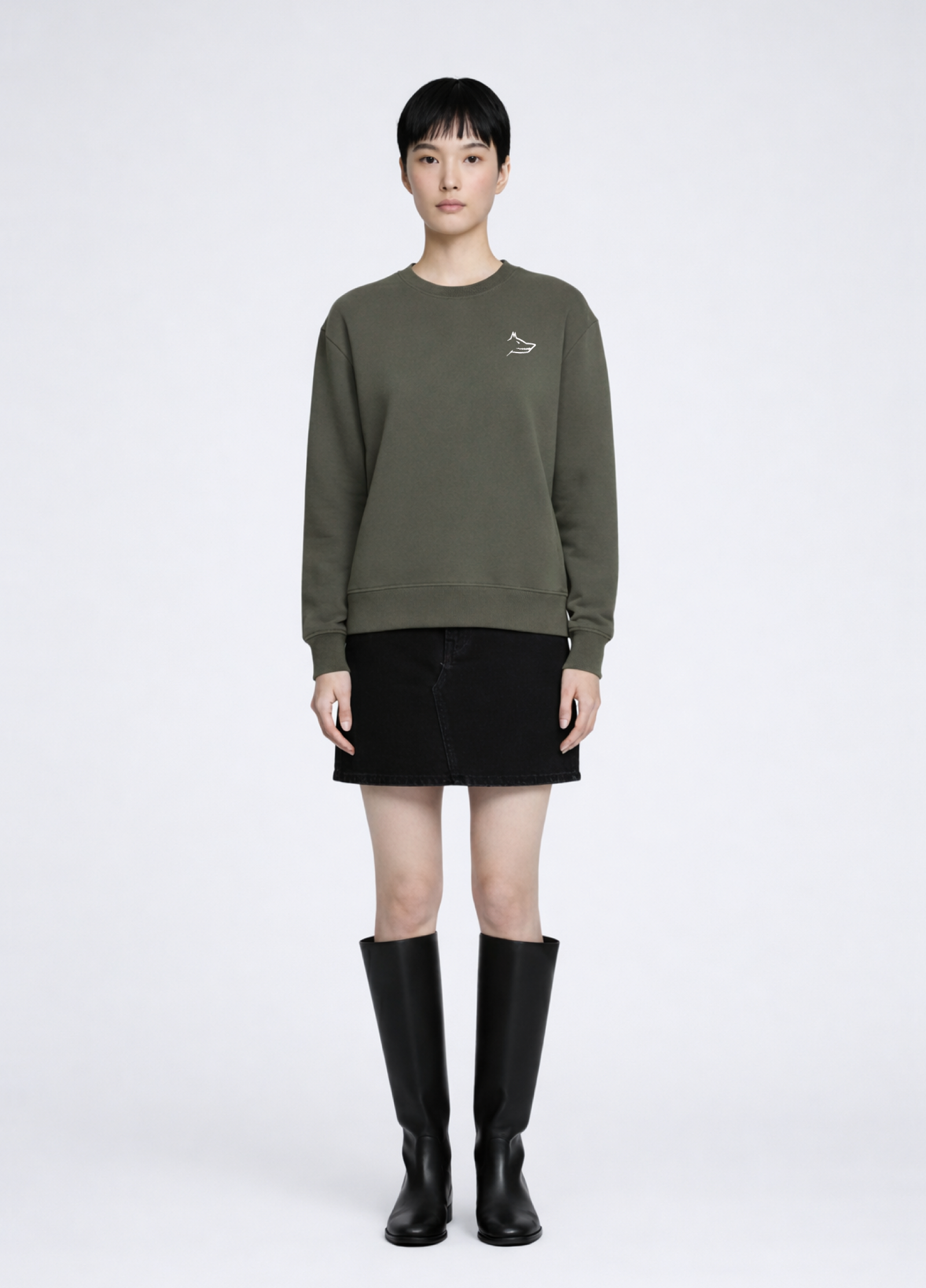 Asian woman wearing a khaki green Northwind Crossword sweatshirt with Northwind wolf embroidered logo on the chest, black skirt, and black knee-high boots on a white background