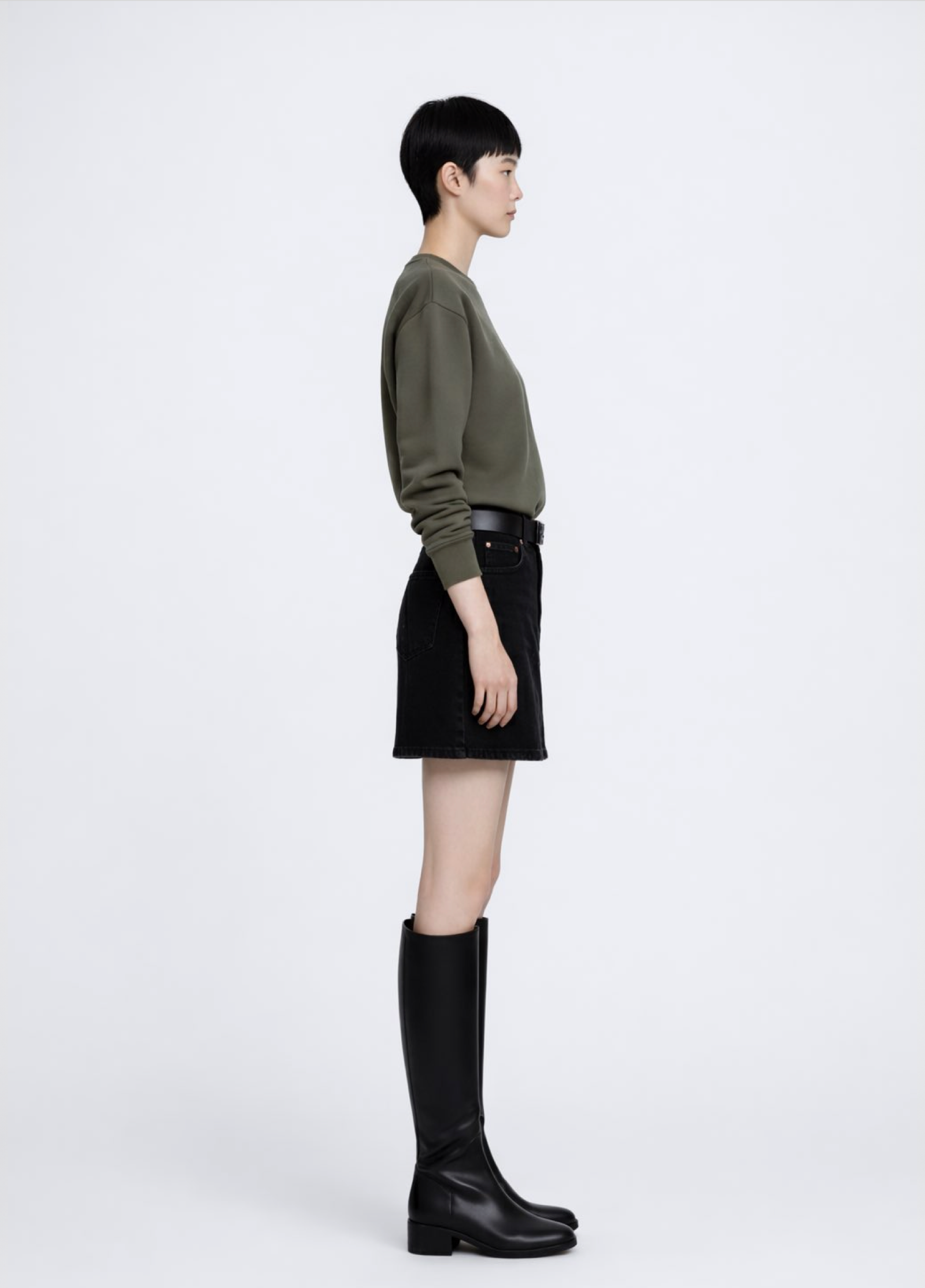 Asian woman wearing a khaki green Northwind Crossword sweatshirt, black skirt, and black knee-high boots on a white background