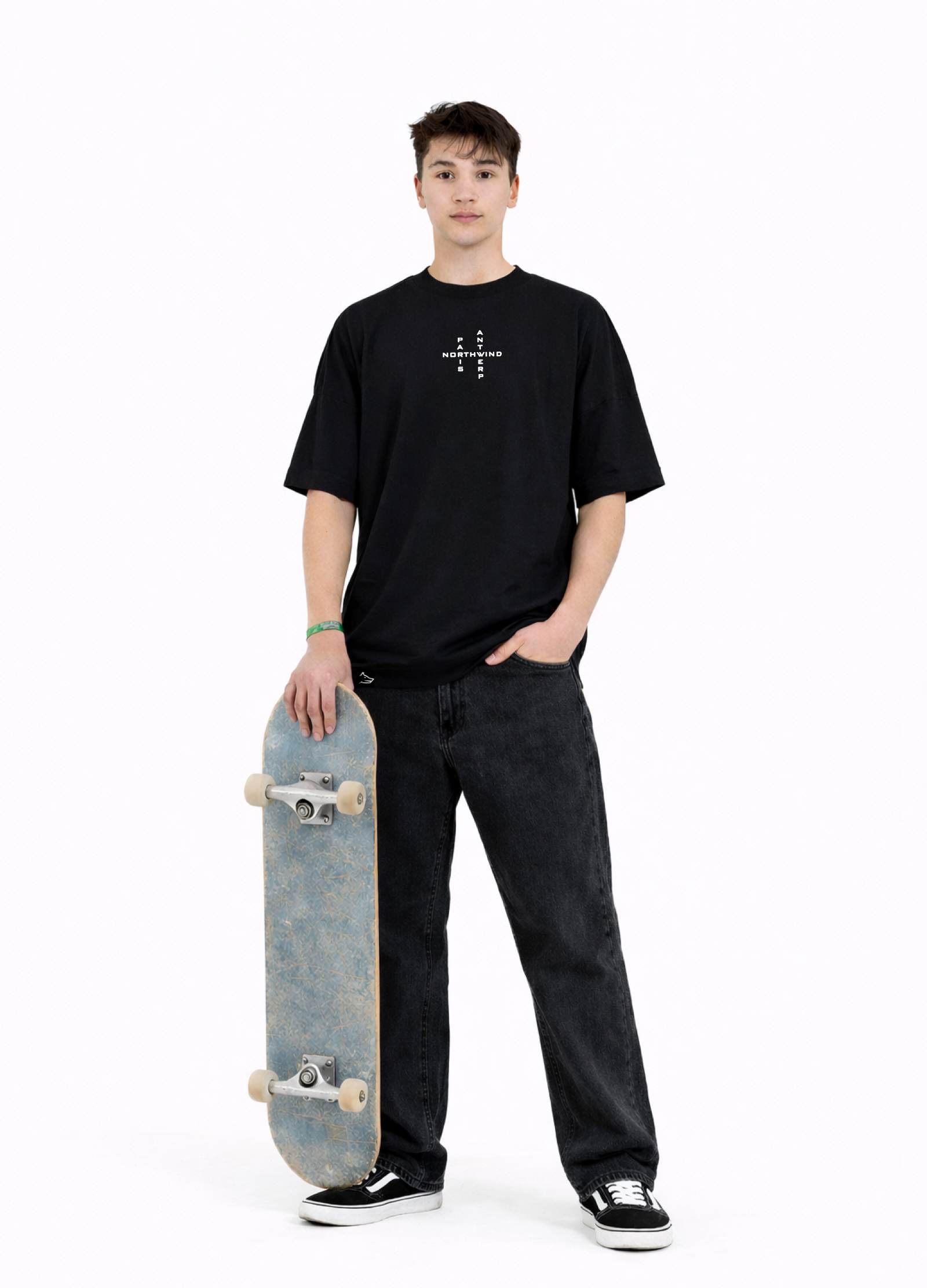 Young adult wearing a Northwind oversized organic cotton black t-shirt with Northwind crossword ANTWERP and PARIS logo embroidered on the chest, Carhartt pants, black Vans shoes, holding a skate board