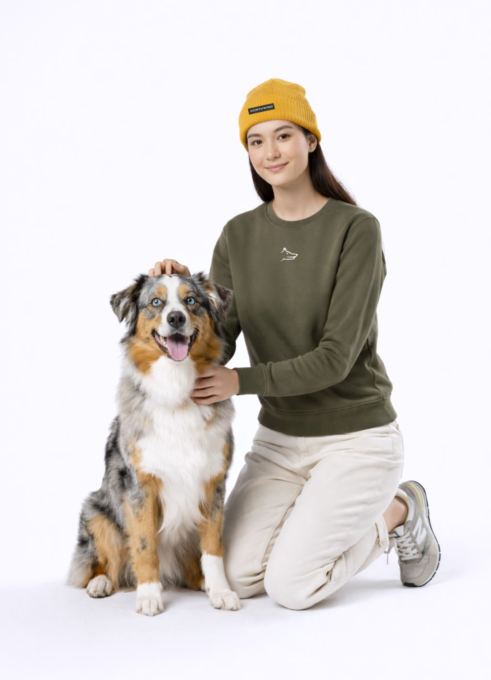 A woman wearing a khaki green Northwind Lone wolf sweatshirt with Northwind wolf embroidered logo on the chest, Northwind organic mustard beanie with a Australian Shepherd dog