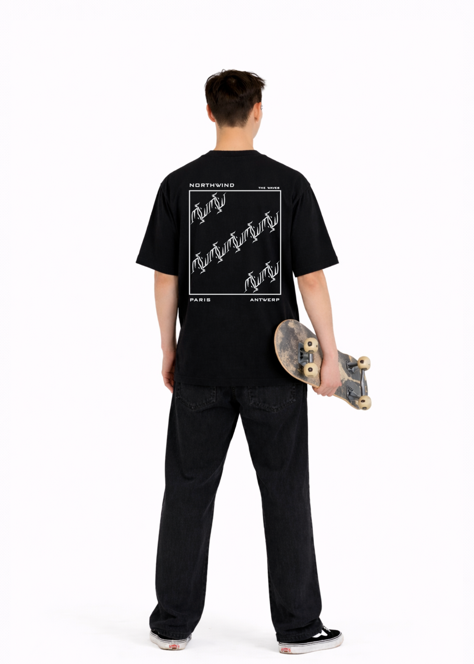Young adult wearing a Northwind oversized organic cotton black t-shirt with Northwind embroidered on the chest, the waves design screen printed on the back, Carhartt pants, black Vans shoes, holding a skate board