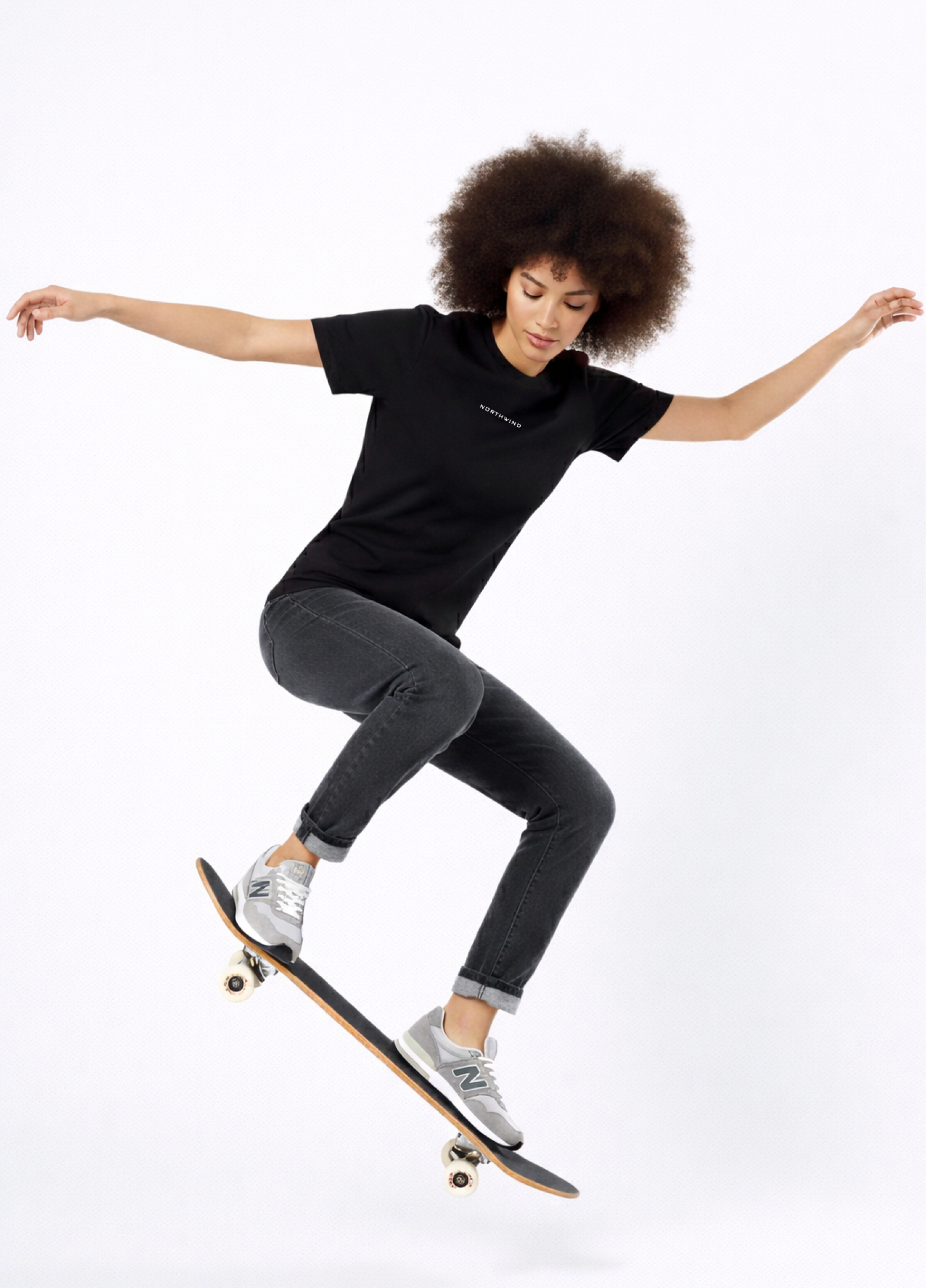 Woman wearing a black Northwind casual t-shirt with minimalist Northwind embroidered logo on the chest and Northwind wolf logo at the back, skateboarding on a white background