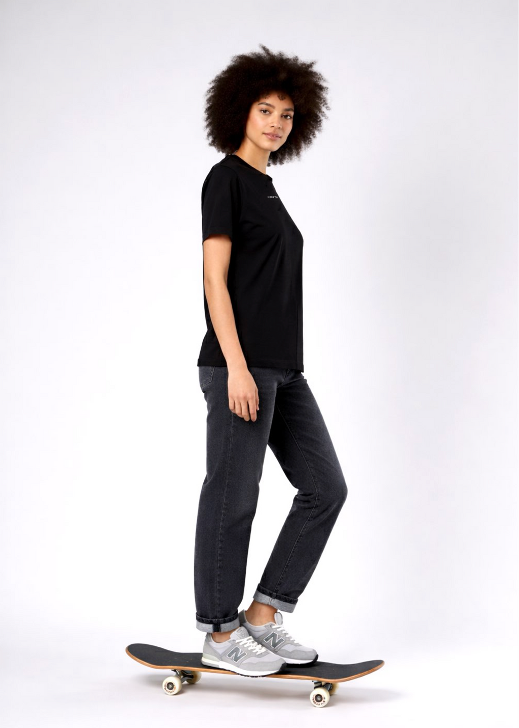 Woman standing on a skateboard wearing a black Northwind casual t-shirt with minimalist Northwind embroidered logo on the chest, gray pants and new balance sport shoes