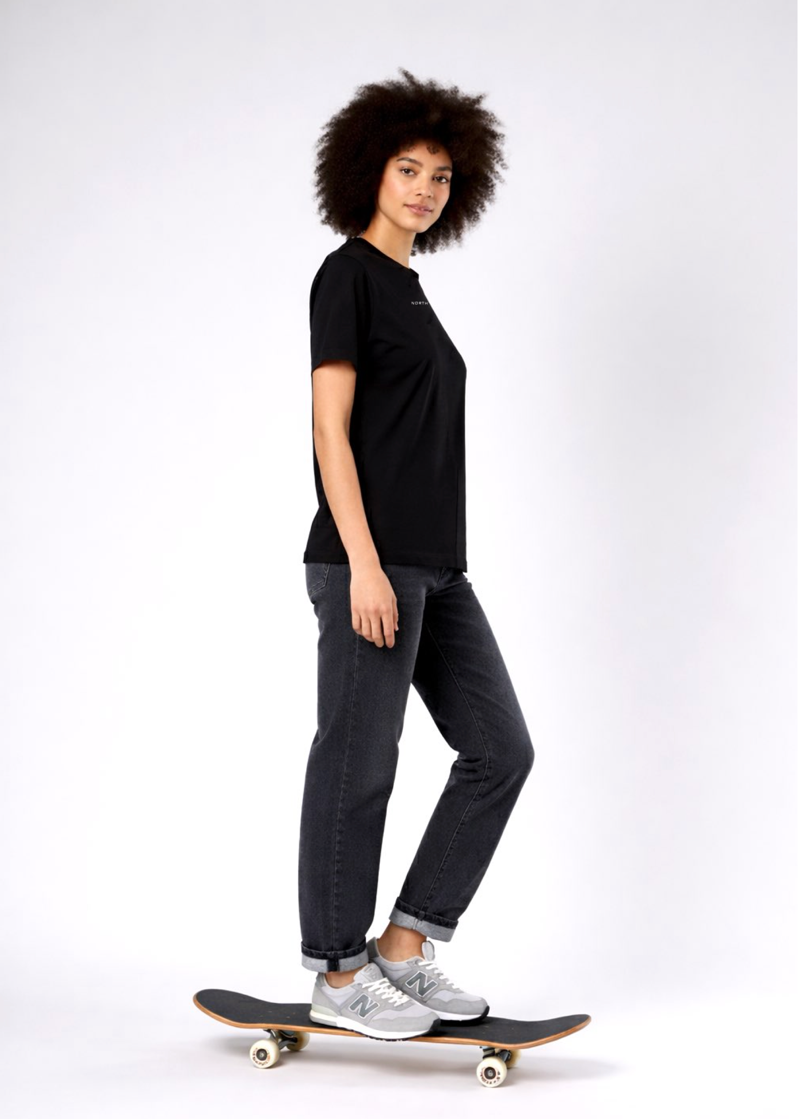 Woman standing on a skateboard wearing a black Northwind casual t-shirt with minimalist Northwind embroidered logo on the chest, gray pants and new balance sport shoes