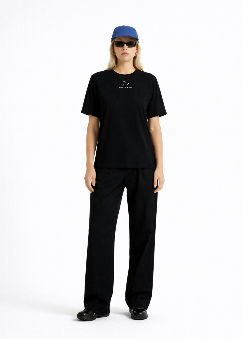 Woman wearing a black Northwind casual t-shirt with minimalist Northwind embroidered wolf logo on the chest, blue hat, black sunglasses and black pants on a white background