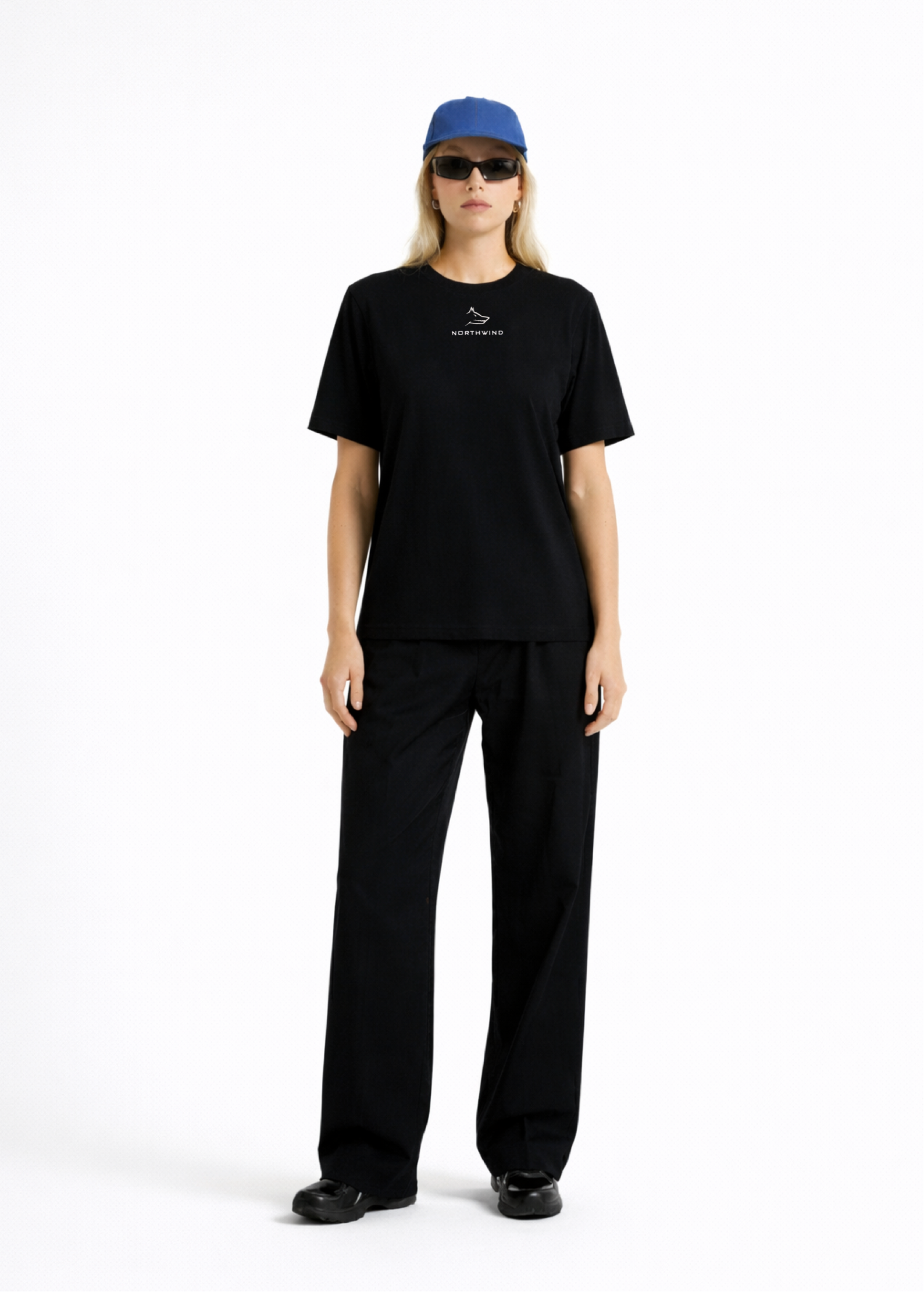 Woman wearing a black Northwind casual t-shirt with minimalist Northwind embroidered wolf logo on the chest, blue hat, black sunglasses and black pants on a white background