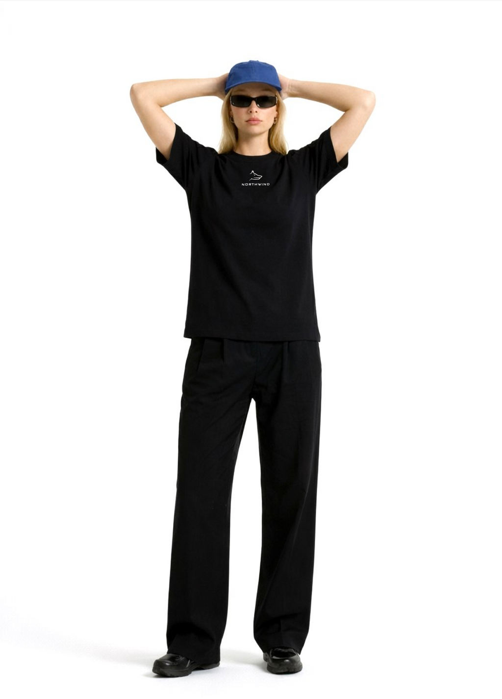 Woman wearing a black Northwind casual t-shirt with minimalist Northwind embroidered wolf logo on the chest, blue hat, black sunglasses and black pants, posing