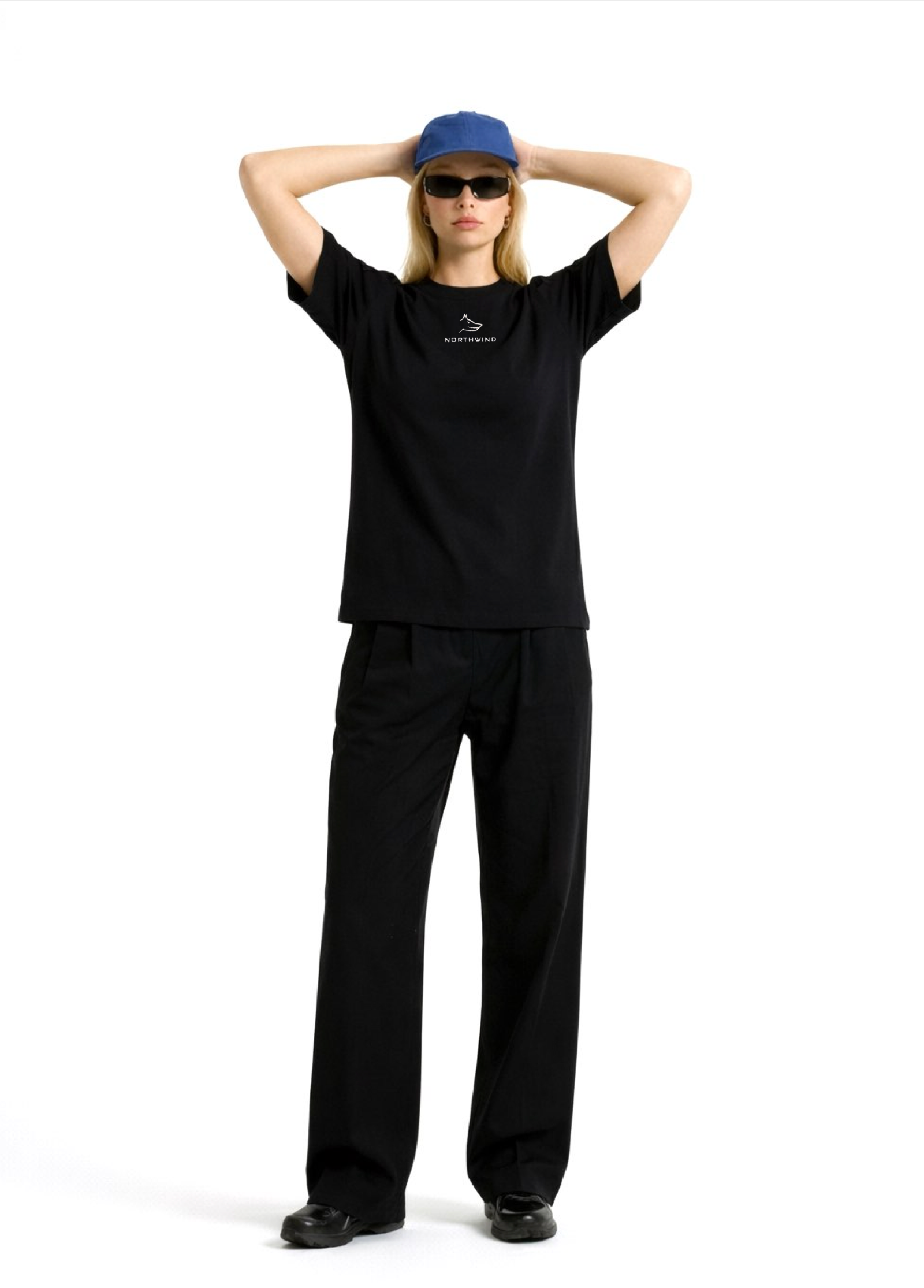 Woman wearing a black Northwind casual t-shirt with minimalist Northwind embroidered wolf logo on the chest, blue hat, black sunglasses and black pants, posing