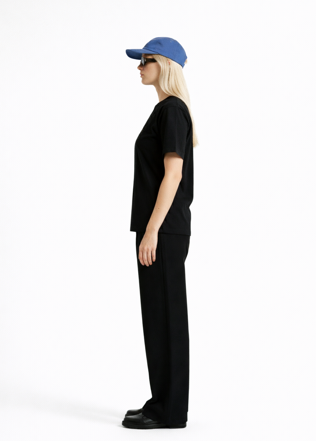 Woman wearing a black Northwind casual t-shirt with minimalist Northwind embroidered wolf logo on the chest, blue hat, black sunglasses and black pants, side pose