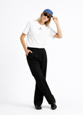 Blond woman wearing a white Northwind casual t-shirt with minimalist Northwind embroidered wolf logo on the chest tucked ın her black pants, blue hat, black sunglasses