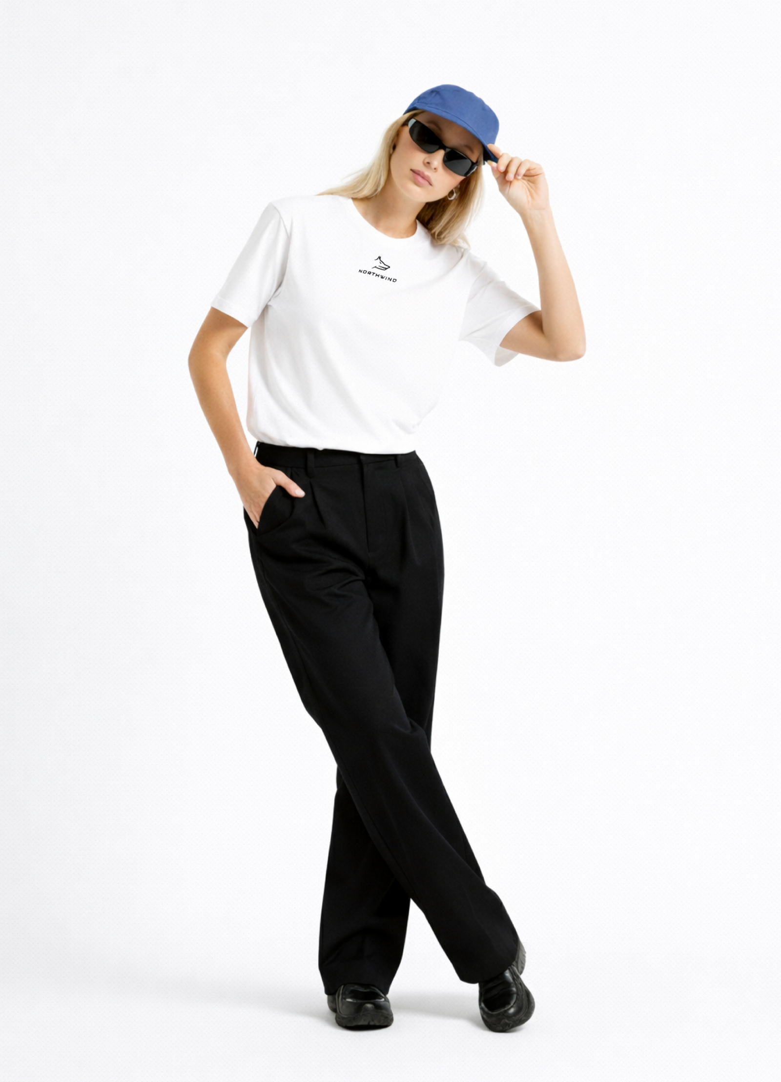 Blond woman wearing a white Northwind casual t-shirt with minimalist Northwind embroidered wolf logo on the chest tucked ın her black pants, blue hat, black sunglasses