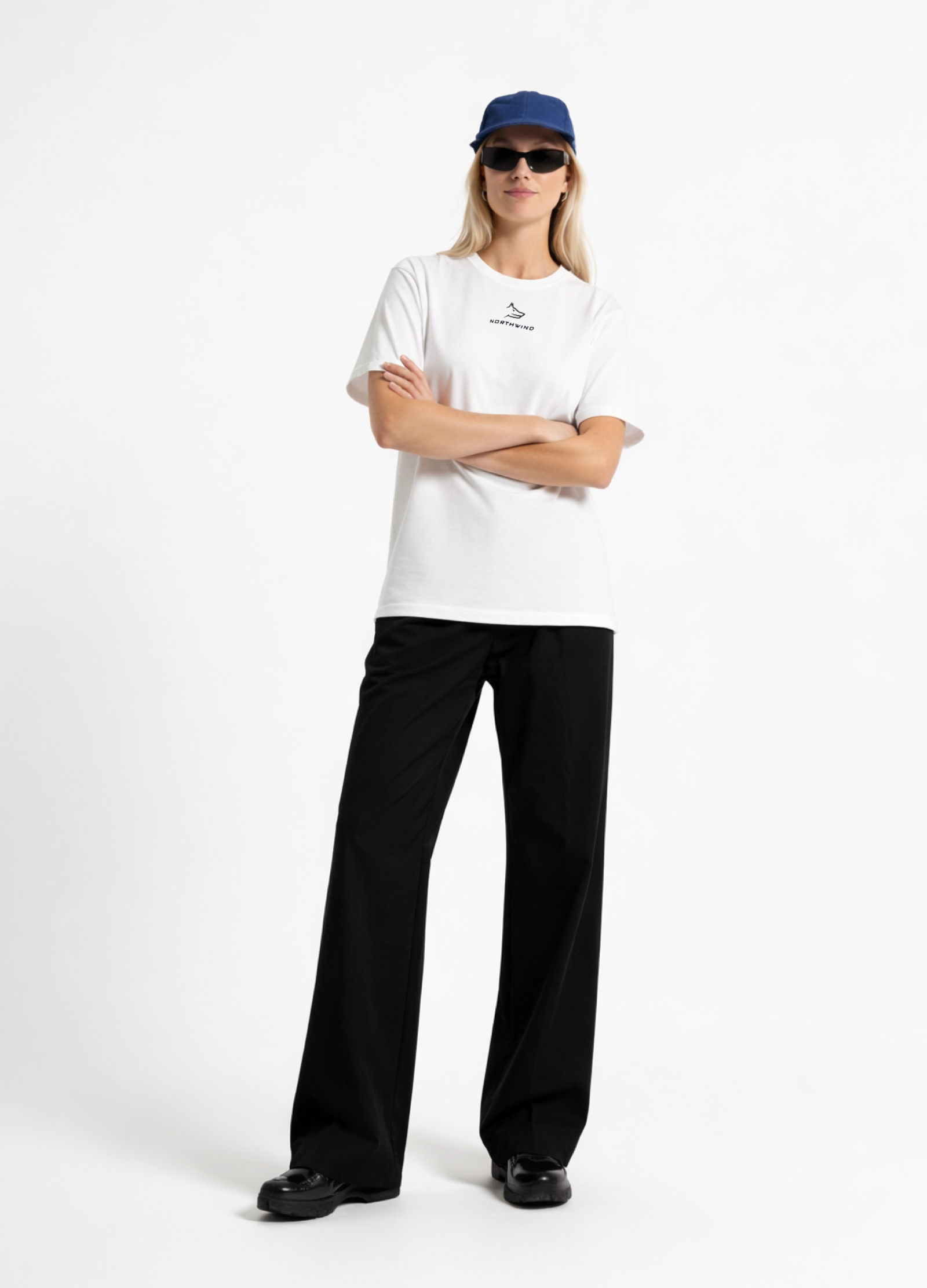 Woman wearing a white Northwind casual t-shirt with minimalist Northwind embroidered wolf logo on the chest, blue hat, black sunglasses and black pants on a white background