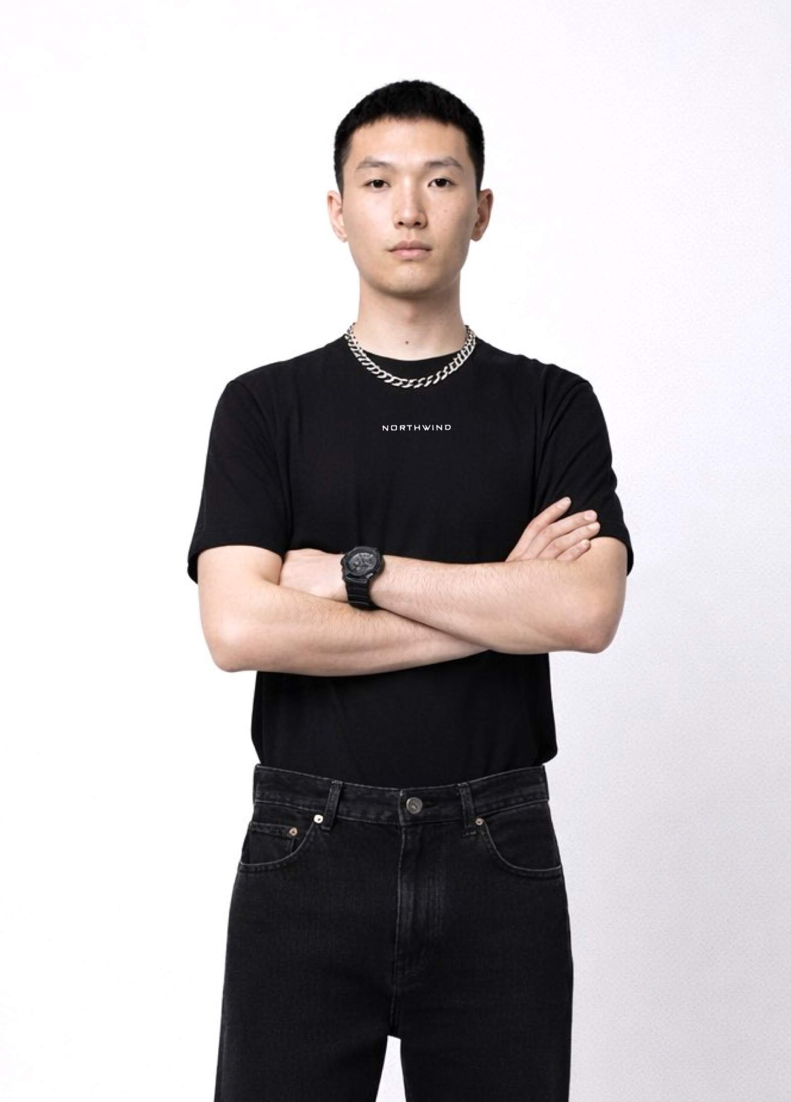Man wearing an essential Northwind black t-shirt with minimalist Northwind embroidered Northwind logo on the chest, Dr. Martens boots, Rolex watch, Carhartt black pants with arms crossed on a white background