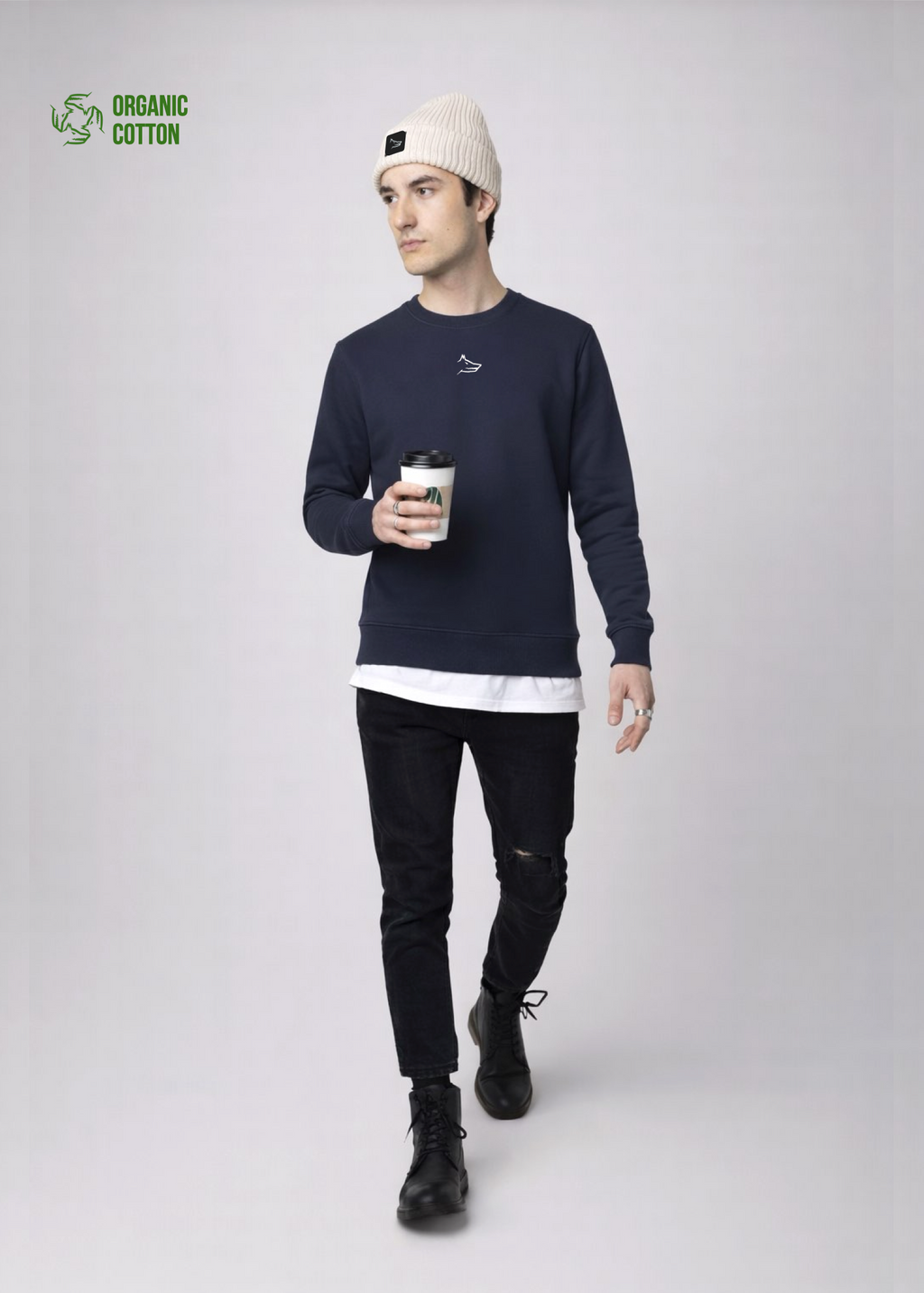 Person wearing a navy blue Northwind sweatshirt with white embroidered Northwind wolf logo on the chest, black jeans, and a sand cream organic beanie with Northwind wolf logo on it and holding a coffee cup on a light gray background