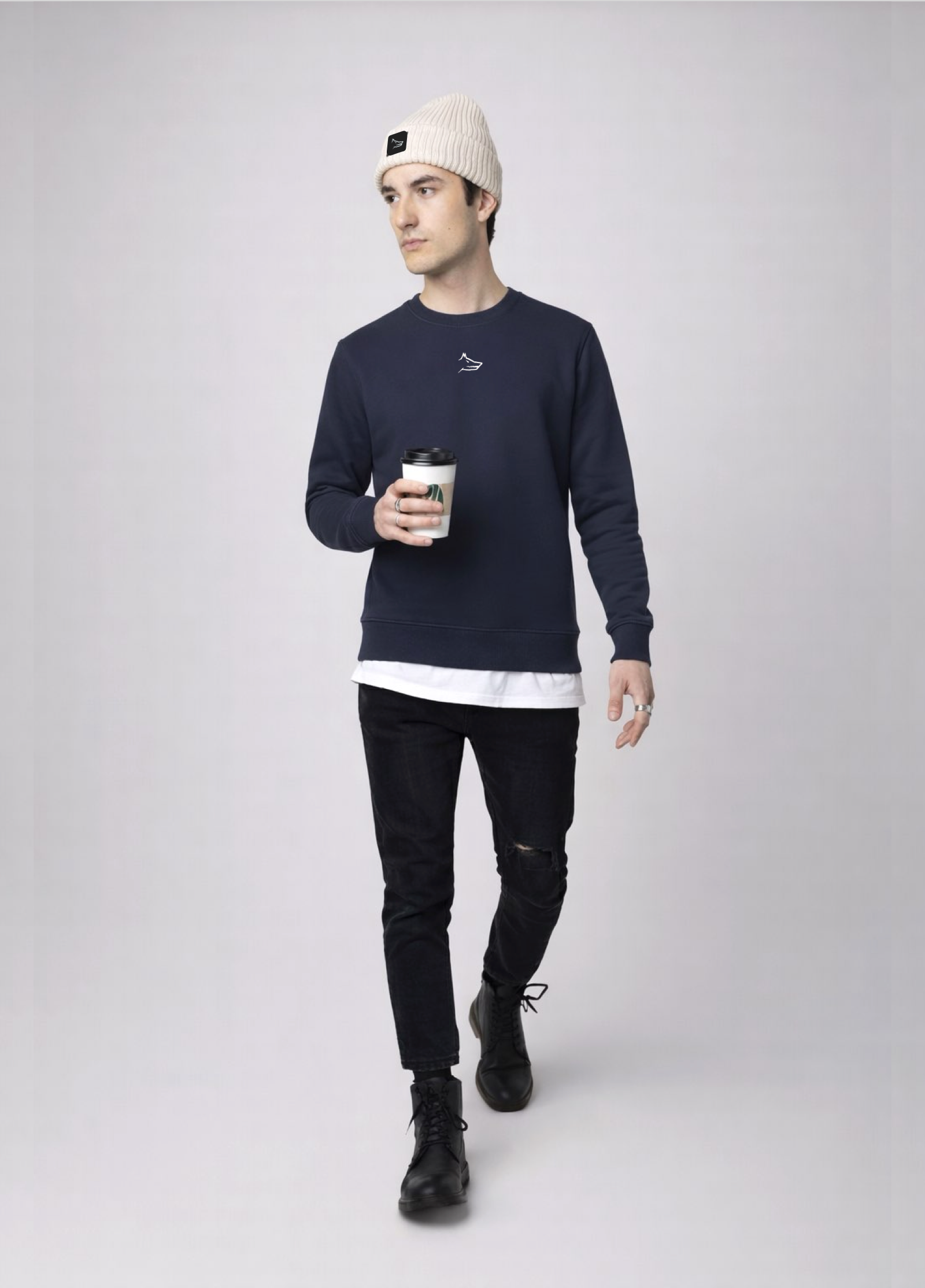 Person wearing a navy blue Northwind sweatshirt with white embroidered Northwind wolf logo on the chest, black jeans, and a sand cream organic beanie with Northwind wolf logo on it and holding a coffee cup on a light gray background