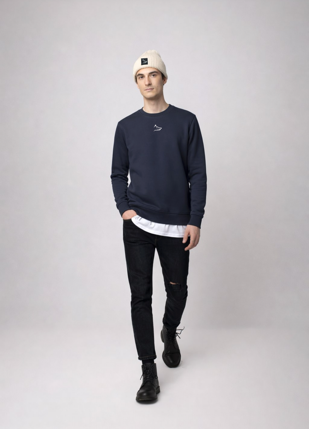 Person wearing a navy blue Northwind sweatshirt with white embroidered Northwind wolf logo on the chest, black jeans, and a sand cream organic beanie with Northwind wolf logo on it