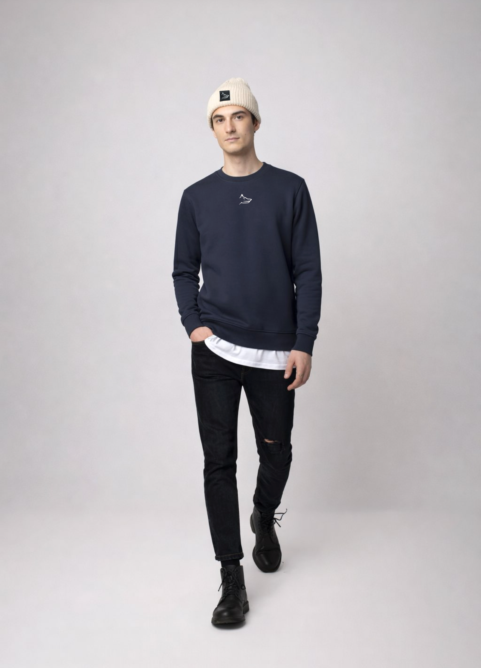 Person wearing a navy blue Northwind sweatshirt with white embroidered Northwind wolf logo on the chest, black jeans, and a sand cream organic beanie with Northwind wolf logo on it
