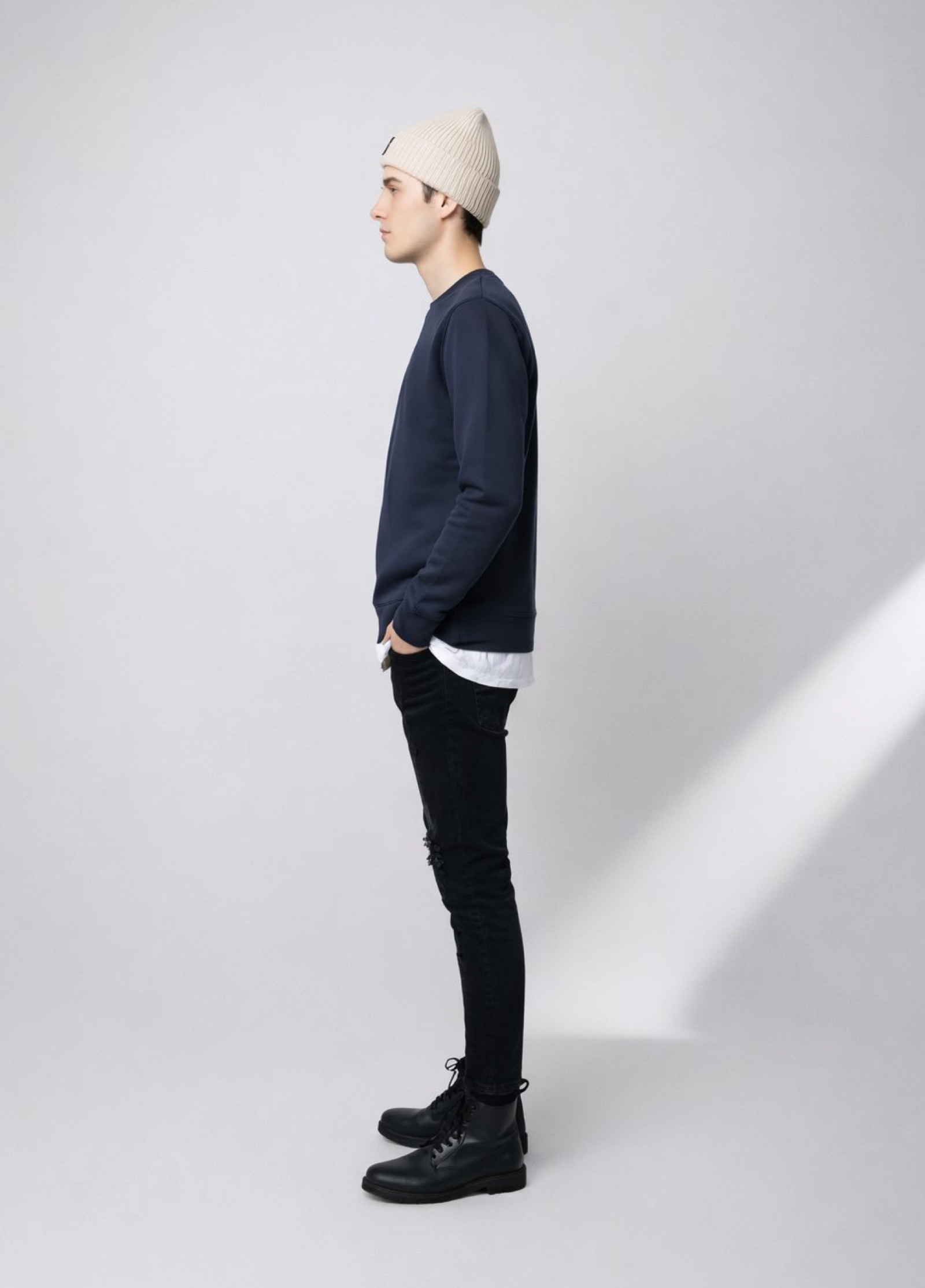 Person wearing a navy blue Northwind sweatshirt with white embroidered Northwind wolf logo on the chest, black jeans, and a sand cream organic beanie with Northwind wolf logo on it, side pose