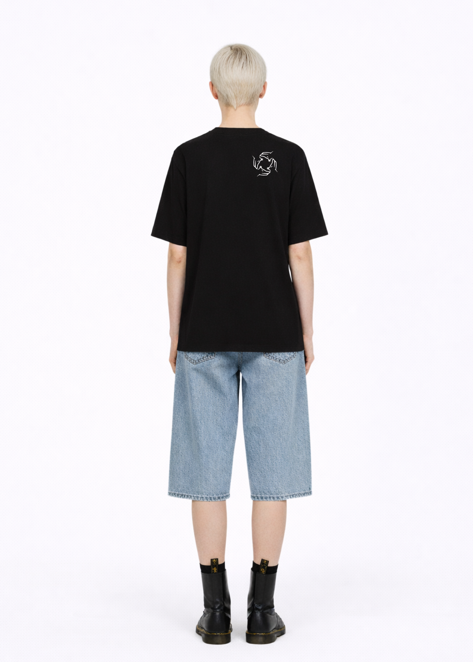 Blond, short hair girl wearing a black Northwind casual storm of wolves t-shirt with Northwind storm of wolves logo embroidered on the back, blue denim Carhartt oversized shorts, black belt, and black Dr. Martens boots