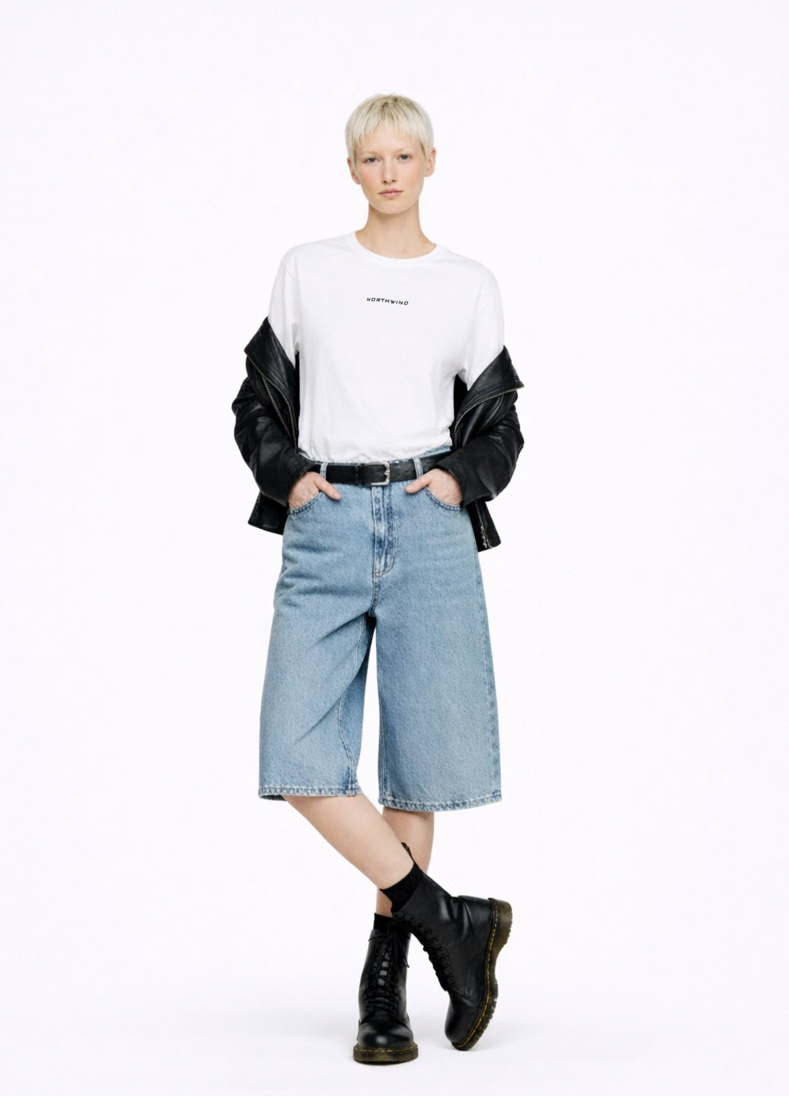Blond, short hair girl wearing a white Northwind casual storm of wolves t-shirt with Northwind logo embroidered on the chest, blue denim Carhartt oversized shorts, black belt, and black Dr. Martens boots on a white background