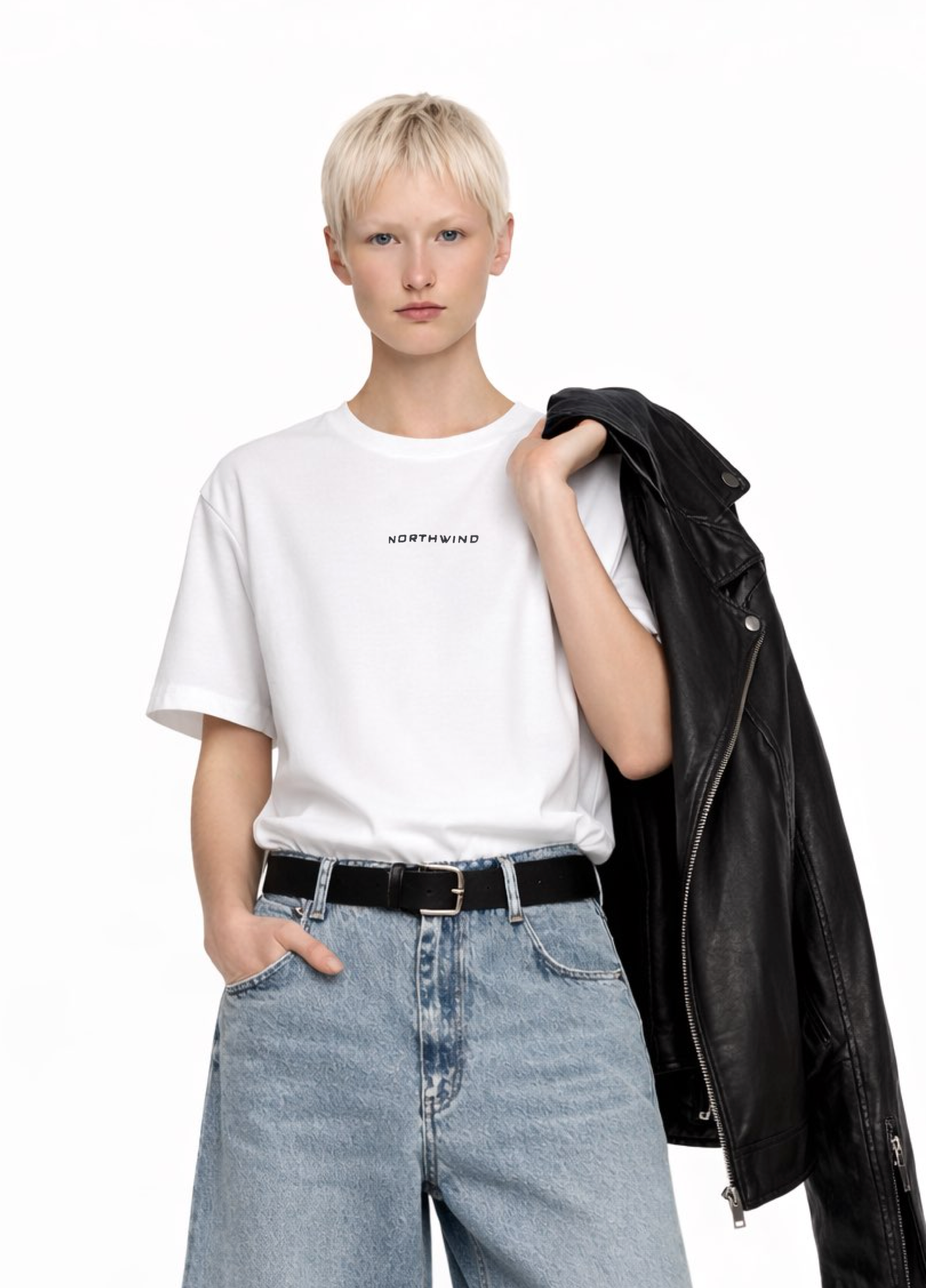 Blond, short hair girl wearing a white Northwind casual storm of wolves t-shirt with Northwind logo embroidered on the chest, blue denim Carhartt oversized shorts, black belt, holding a black leather jacket.