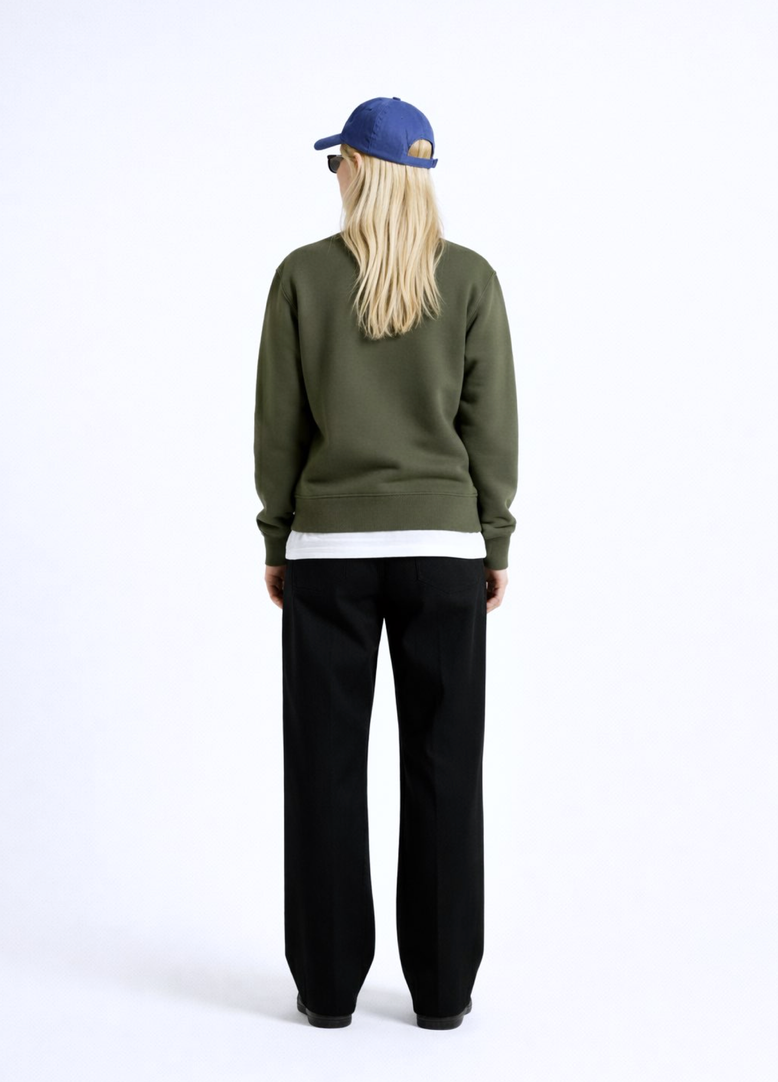 Woman wearing an olive khaki green Northwind sweatshirt with Northwind storm of wolves embroidered logo on the chest, black pants, and a blue cap on a white background