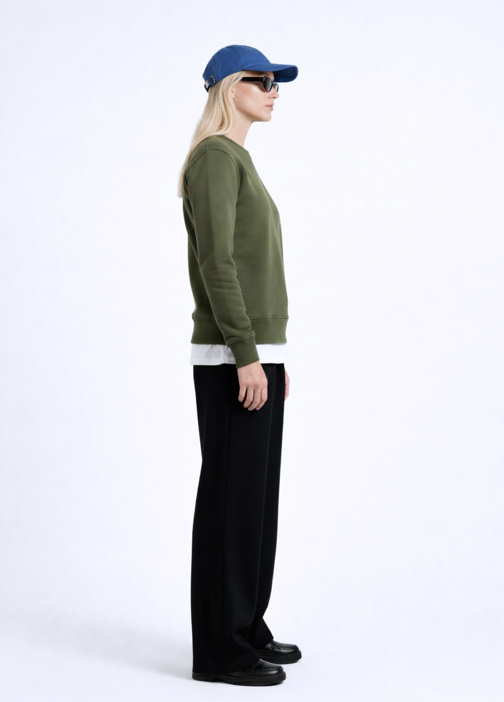 Woman wearing an olive khaki green Northwind sweatshirt with Northwind storm of wolves embroidered logo on the chest, black pants, and a blue cap on a white background