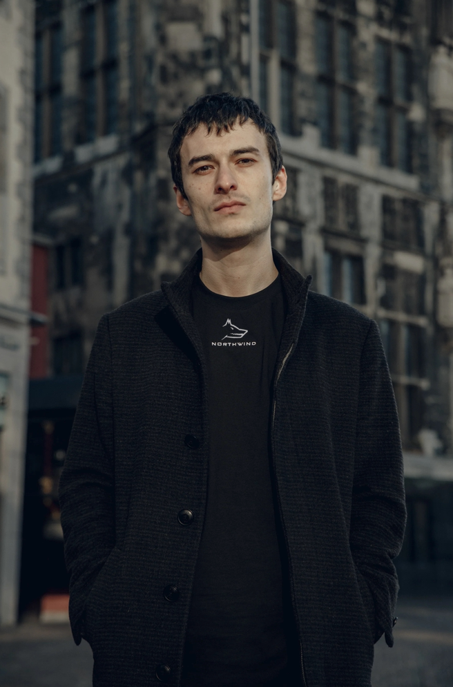 Man wearing a black coat and Northwind black t-shirt with a logo, standing in an urban setting.