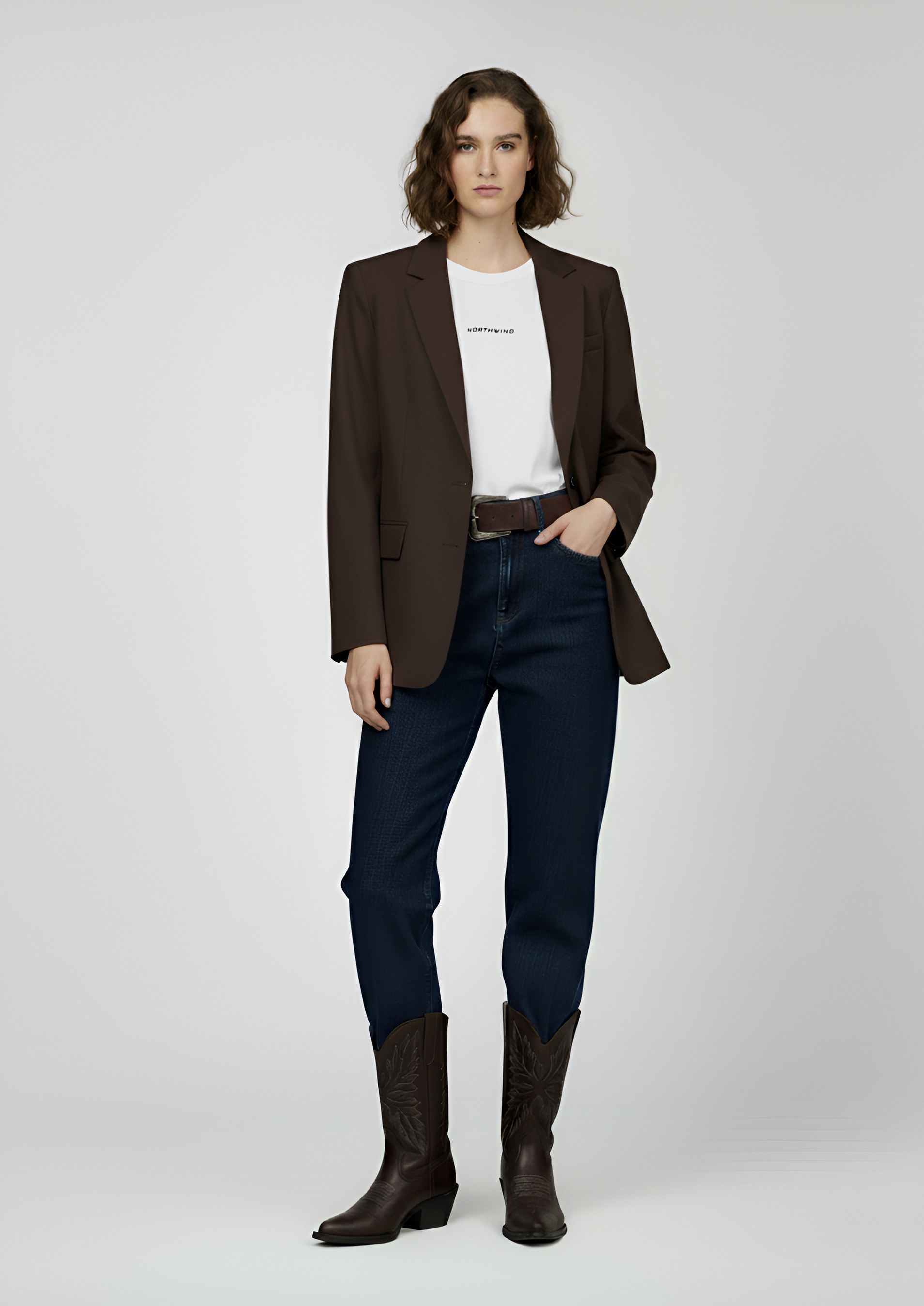 Woman wearing a brown blazer, white Northwind t-shirt, dark blue jeans, and brown boots on a white background