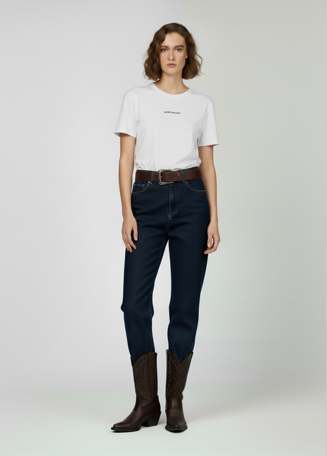 Woman wearing a white Northwind t-shirt, dark jeans, and brown boots on a plain background
