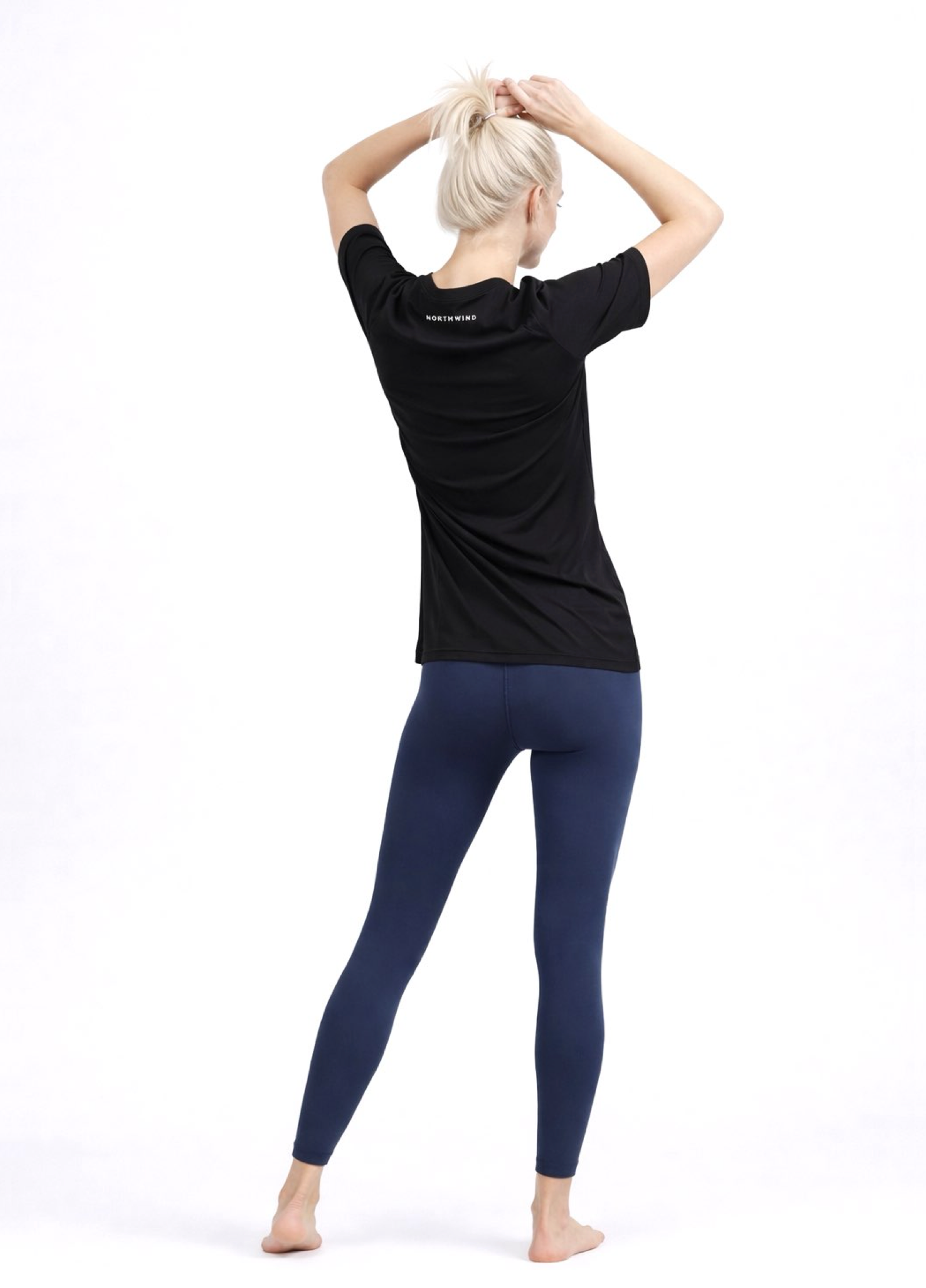 Woman wearing a black Northwind activewear t-shirt with 'NORTHWIND' branding on the back, blue legging, standing against a white background.
