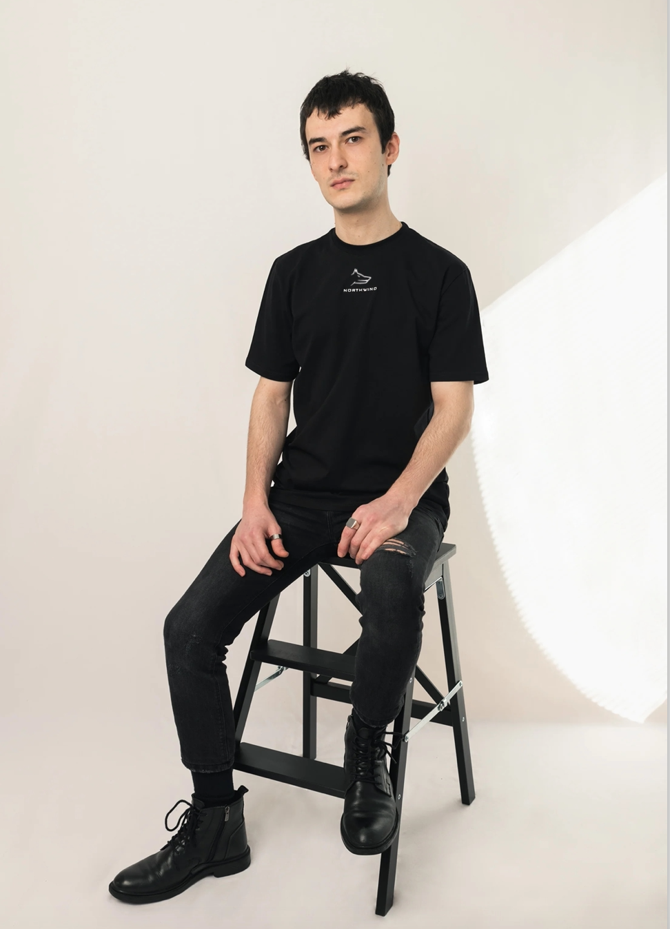 Man sitting on a stool wearing a Northwind casual essential black t-shirt and pants against a white background,  Northwind embroidery wolf logo on a light background