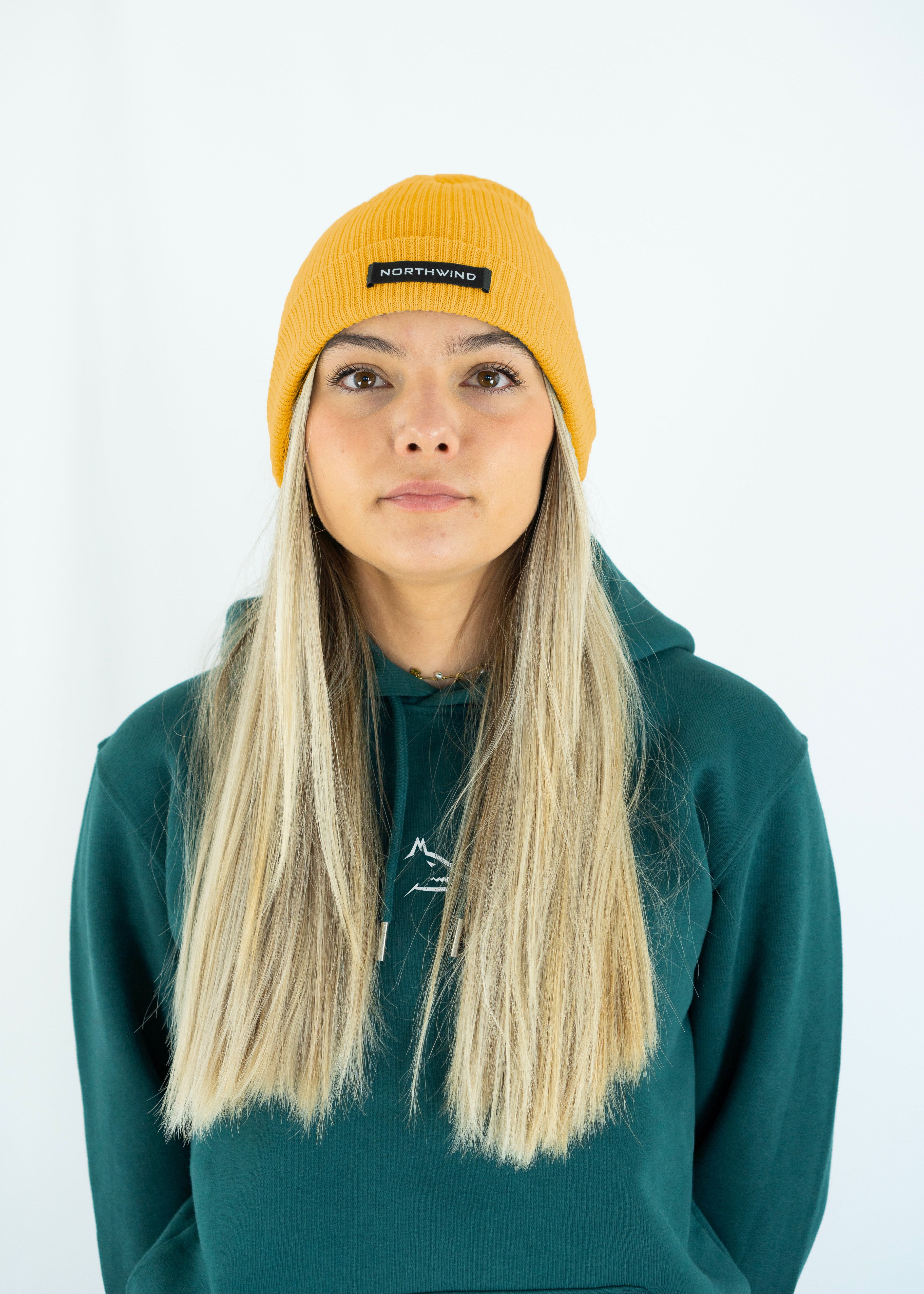 Woman wearing a mustard organic cotton beanie with a Northwind brand logo, and a green Northwind hoodie on a white background