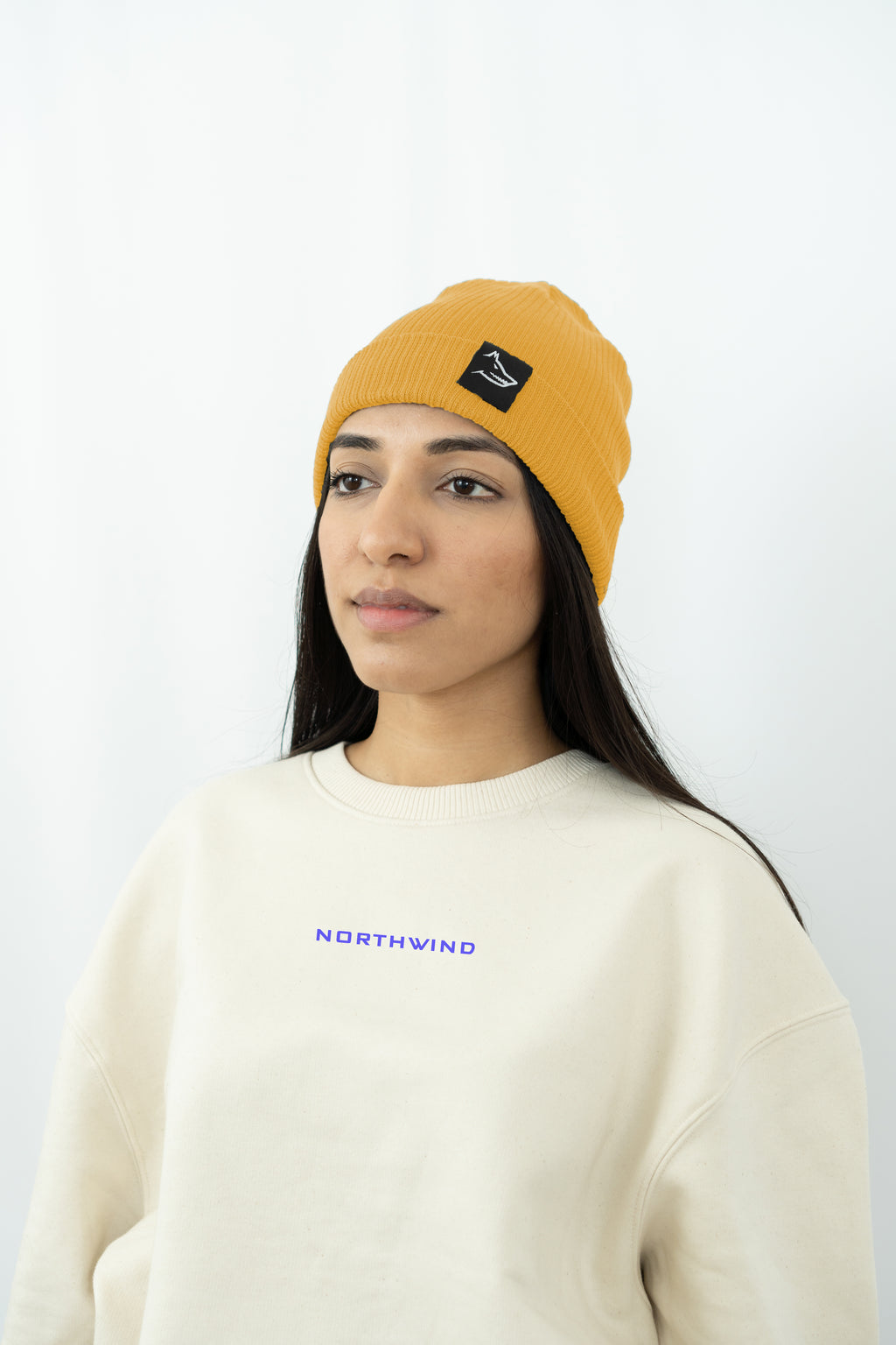 woman wearing a mustard organic cotton beanie with a Northwind wolf logo, and a beige oversized Northwind organic cotton sweatshirt with Northwind logo on the chest